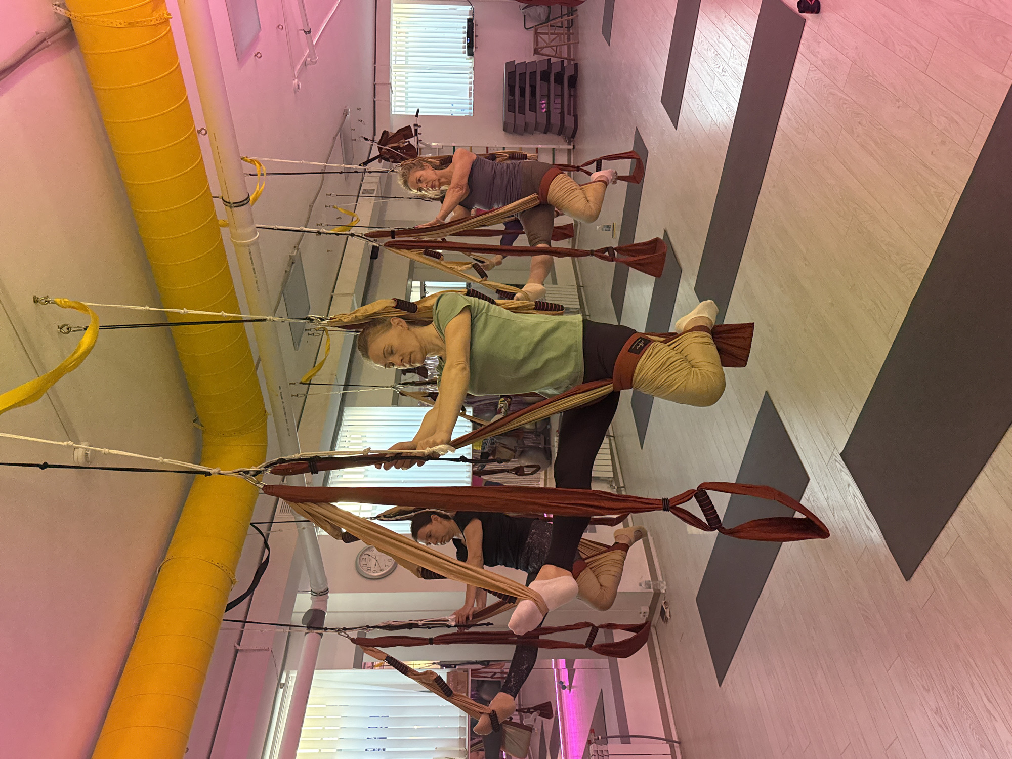 The image shows an indoor space, possibly a yoga or fitness studio, with three individuals suspended in aerial yoga hammocks. The individuals are performing poses, each with one leg extended and the other bent.
The environment features a pink-toned ceiling with yellow ductwork and supporting cables for the hammocks. A wall with a window and blinds is visible in the background, along with shelving and equipment. The floor is light wood, marked with dark gray geometric shapes. A clock is visible on the wall in the middle ground.
The image was taken in a fitness studio. Further information on the precise location in Chaikovsky, Russia, cannot be determined from the image.
