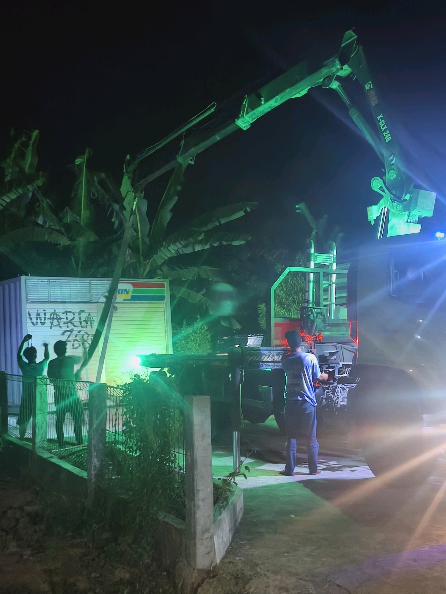 In the city of Kroya, Indonesia, at night, a scene unfolds near a white, corrugated metal structure, possibly a shop or container, with "WARGA" (meaning "residents" or "citizens") spray-painted on it in large letters, along with other markings. Two figures, silhouetted against a bright green light emanating from the structure, stand behind a metal fence. One figure appears to be holding up an object.
Dominating the upper portion of the image is a large green mechanical crane arm, extending from a truck. The crane arm has the inscription "X-CLX 248" visible on it. A person, wearing a blue t-shirt and dark pants, stands on the ground near the truck, facing away from the viewer and towards the crane. The ground is illuminated by strong green and white lights, casting shadows. The environment appears to be outdoors, with large, dark green leaves from tropical plants visible in the background, suggesting a humid, possibly rural or semi-rural setting within Kroya. The overall atmosphere is dramatic due to the nighttime setting and the vibrant green illumination.