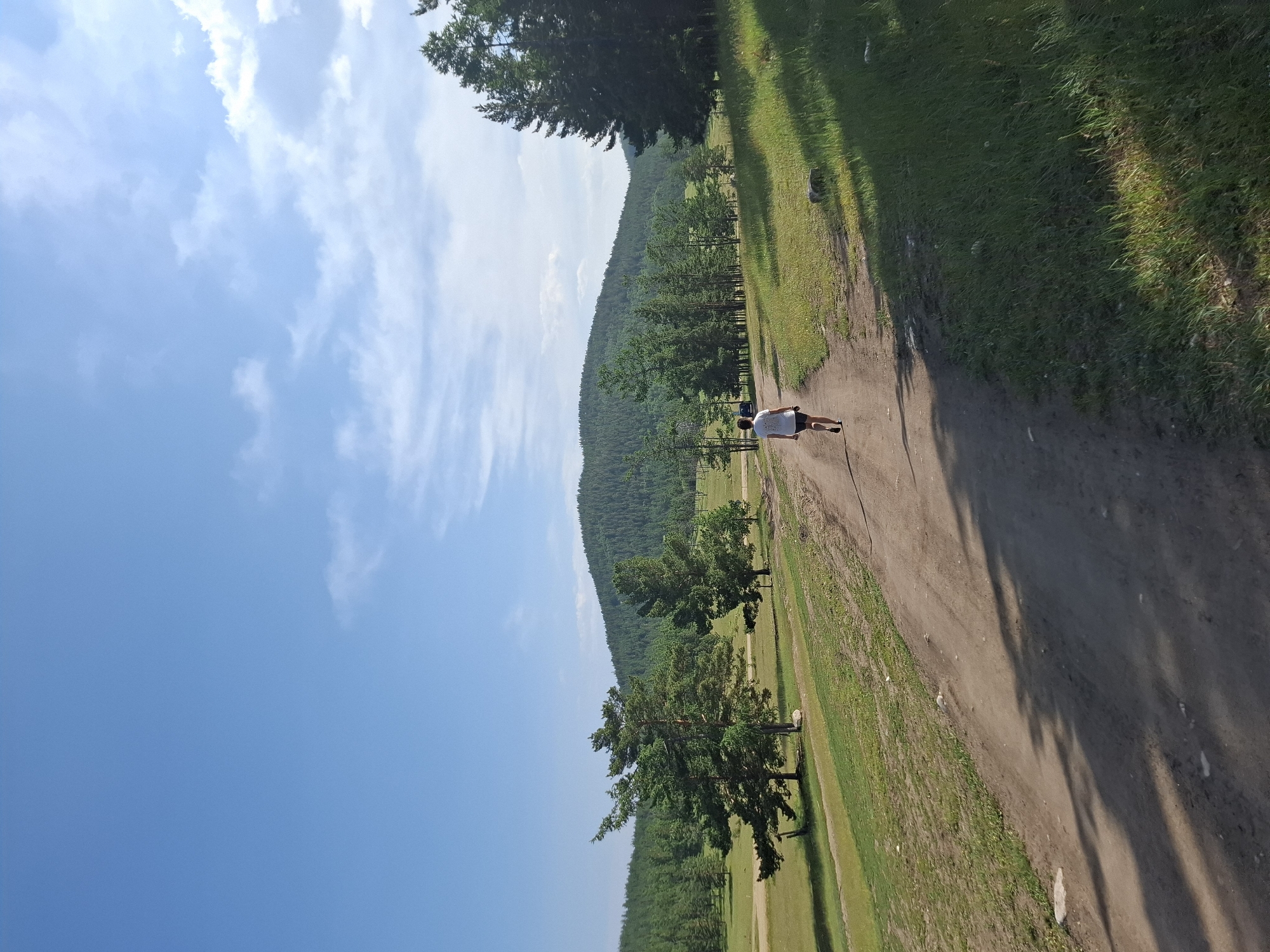 A person walks away from the camera down a dirt path through a grassy landscape. The person is wearing a white t-shirt and dark shorts. To the left of the path, there are several evergreen trees and a low, green hill. In the background, a larger, densely forested mountain rises under a blue sky with scattered white clouds. The scene appears to be a rural outdoor location, possibly in or near Bugul'deika, Russia, a settlement on the shore of Lake Baikal known for its natural beauty and surrounding forests. The lighting suggests it is daytime.