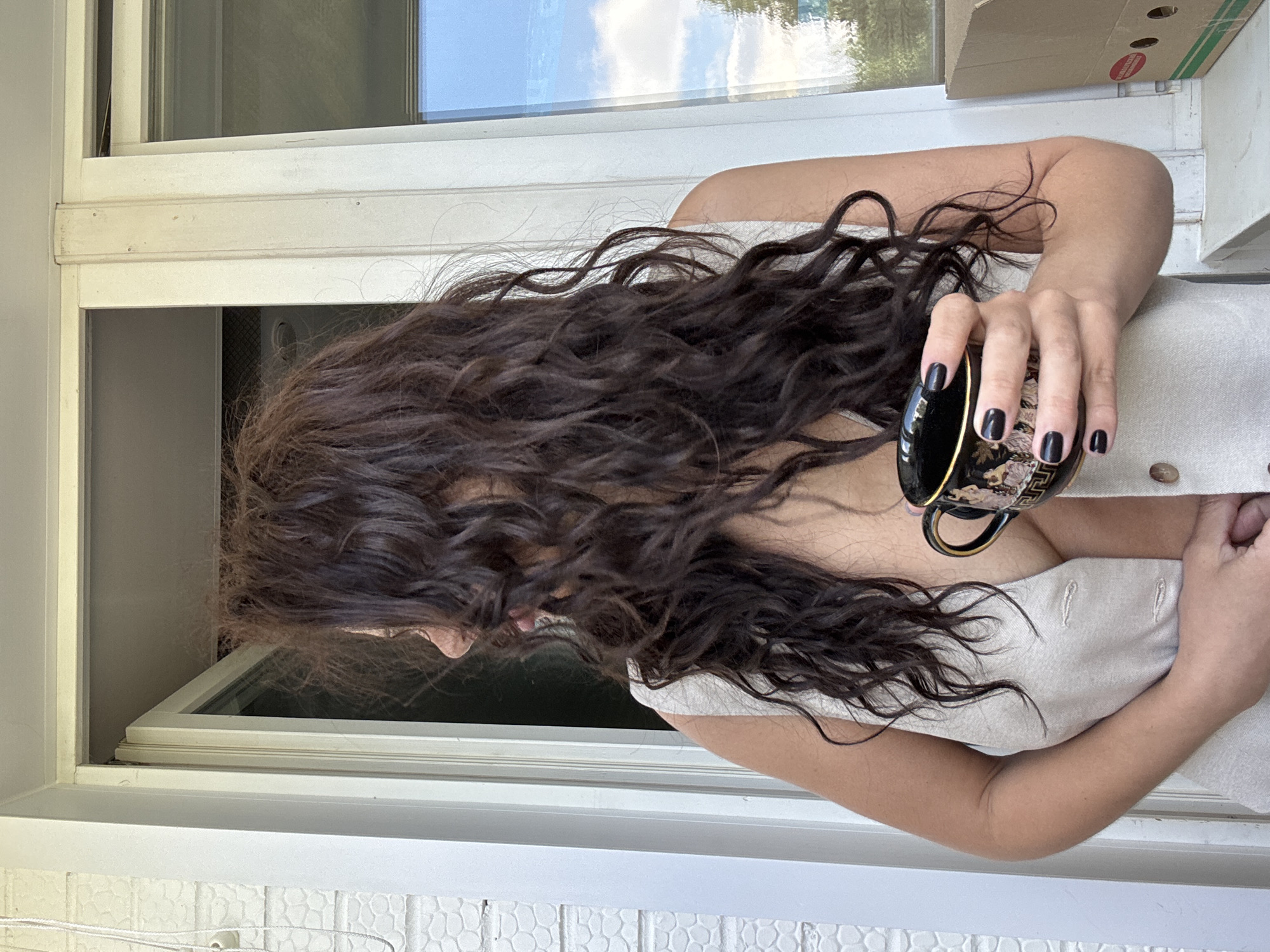 The image shows a person from the chest up, positioned indoors beside a white window frame, oriented towards the left. The subject has long, dark brown, wavy hair that cascades forward, partially obscuring their face. They are wearing a light-colored, possibly beige or off-white, sleeveless garment with visible buttons down the front.
In their right hand, the person holds an ornate black teacup or coffee cup with intricate gold detailing, including what appears to be a Greek key pattern and decorative motifs. Their fingernails are painted with a dark, possibly black, polish. The left hand is also visible, resting gently on the garment near the buttons.
Through the window, a bright sky with white clouds is reflected, indicating it is daytime. Outside the lower portion of the window, a textured white surface, possibly a tiled wall or a railing, is visible. In the upper right background, a cardboard box with an unreadable red and white label is partially seen. The scene suggests a quiet moment, possibly with the person looking out the window or down at their cup. The context provided indicates the location is Chaikovsky, Russia.