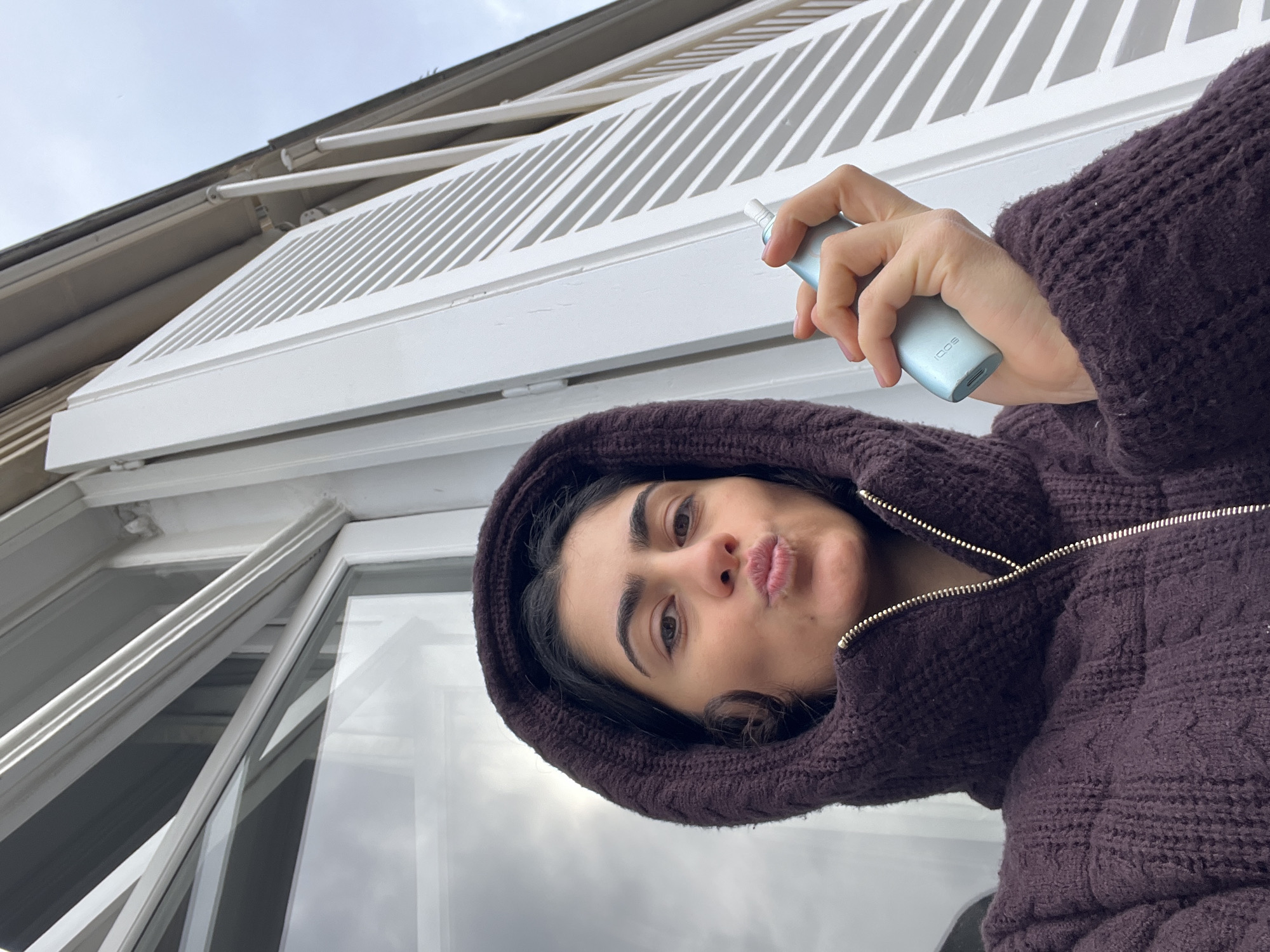 A single adult woman is depicted in a medium shot, posing outdoors against the backdrop of a building in Paris, France. The architectural elements, including a white multi-paned window and a partially open white louvered shutter, are consistent with Parisian structures. The woman has dark hair and brown eyes, looking directly at the camera with her lips puckered. She is wearing a dark purple knitted hooded jacket with a visible silver zipper. In her right hand, she holds a light blue IQOS electronic heated tobacco device, displaying a light pink nail polish on her fingernails. The sky visible through and reflected in the window appears overcast, indicating diffuse daylight. No other individuals are present.