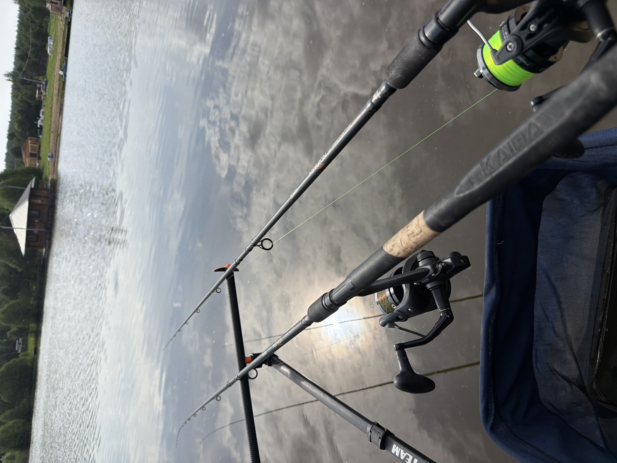 The image shows a close-up view of two fishing rods set up on a rod rest, positioned near a body of water. The rods are equipped with fishing reels. One reel is spooled with bright green line. In the background, a water surface reflects the sky with white cloud patterns. On the left edge of the image, the opposite bank of the water is visible, showcasing trees and a few small buildings. The location is assumed to be Prikamskii, Russia. A blue net is also visible on the right.