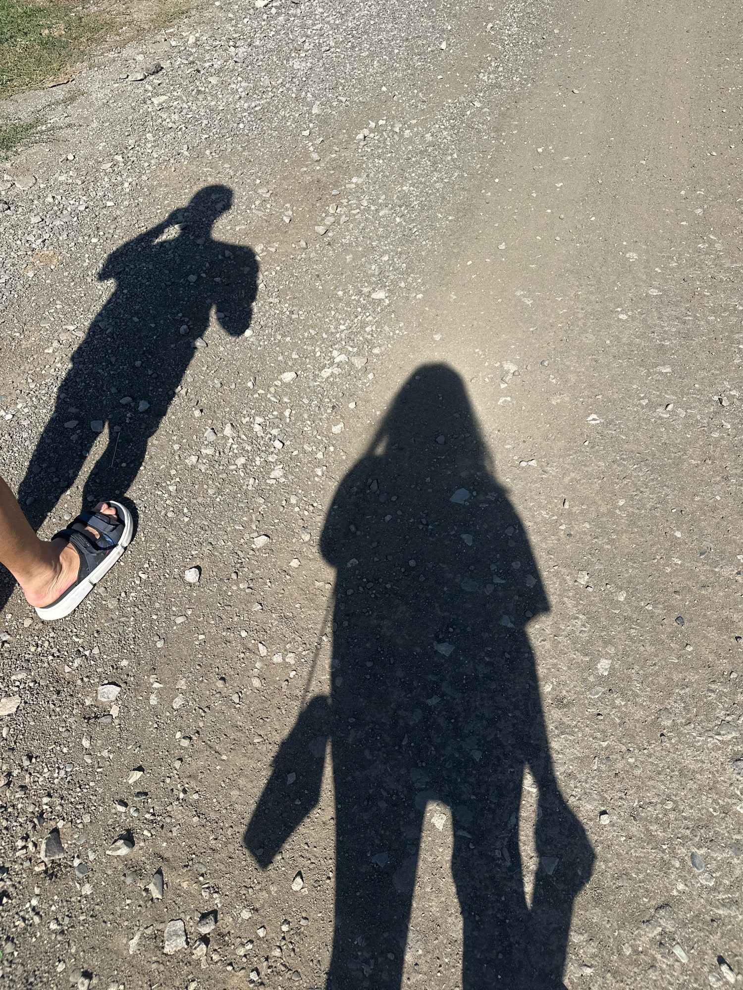 The image shows two shadows cast on a gravelly path. One shadow, on the left, depicts a person holding a phone to their ear. A bare foot wearing a sandal is visible at the bottom left of the frame, belonging to the person casting this shadow. The second, larger shadow, on the right, shows a full figure of a person with a bag hanging from their shoulder. The ground is composed of small stones and dirt. The lighting suggests a sunny day. The scene does not provide any specific visual cues to determine its location in Gyumri, Armenia.