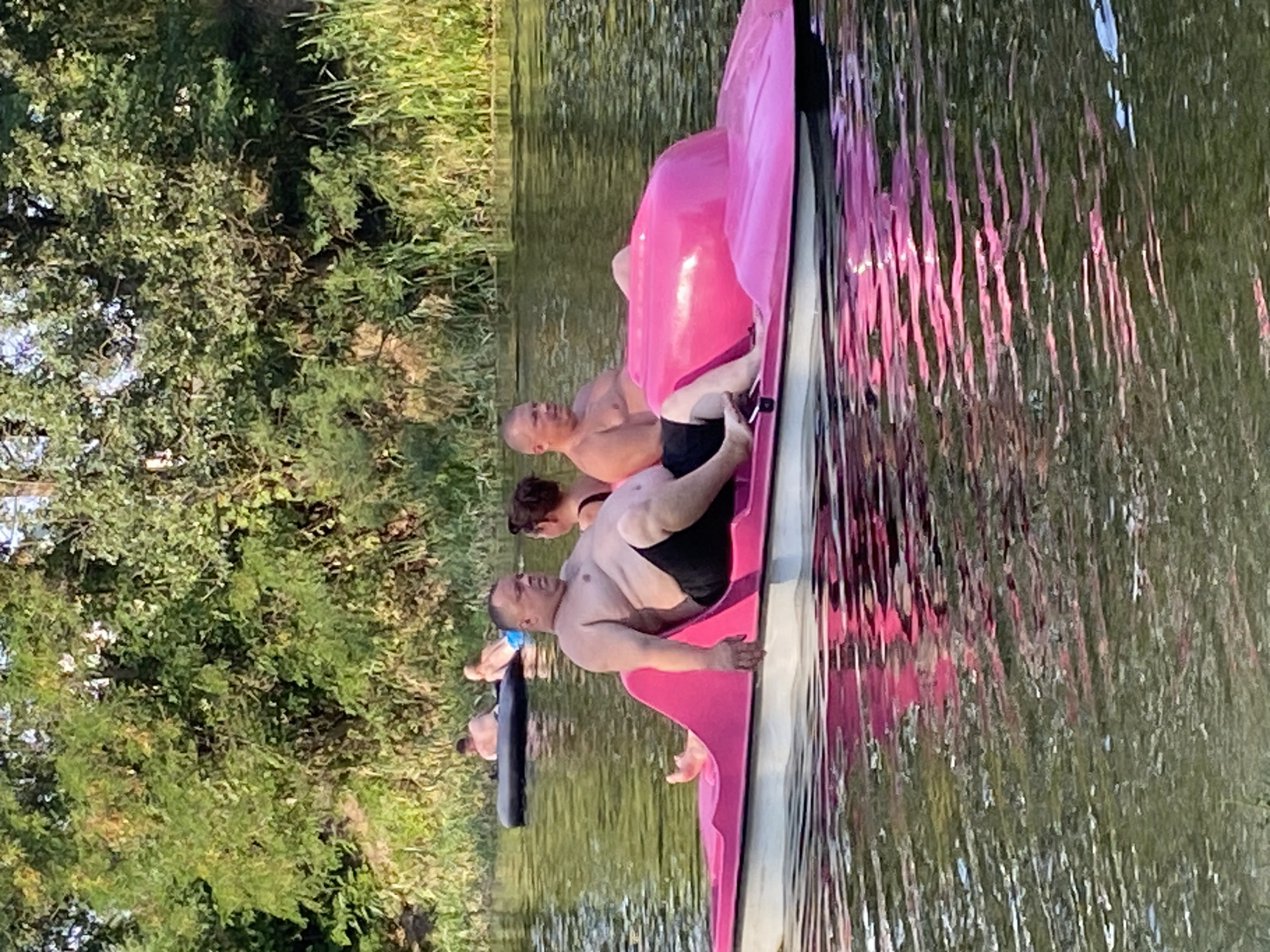 This image depicts a summer day on a body of water, likely a river or lake, with lush green trees and reeds lining the bank in the background. In the foreground, a bright pink pedal boat is occupied by three individuals. A heavyset man with a bare chest and black shorts is seated on the left side of the boat, looking to his right. To his right, a woman with dark hair, wearing a black swimsuit top, is partially visible. On the right side of the boat, a man with a bare chest and a light-colored lower garment is seated, looking towards the left. Further in the background, two men are on an inflatable tube in the water. The water's surface reflects the pink boat and the surrounding greenery. The scene appears to be in Semianivka, Ukraine, suggesting a recreational setting.