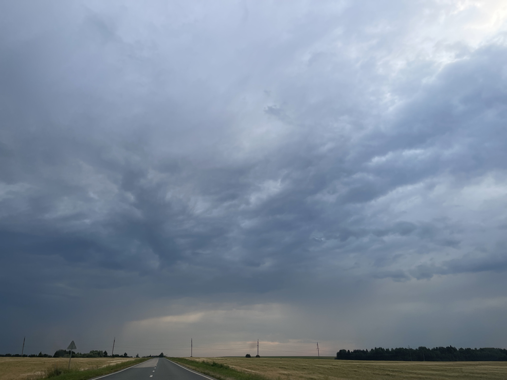 The image depicts a vast, overcast sky filled with heavy, grey clouds, suggesting an impending storm. A paved road stretches into the distance, bordered by fields of dry, golden grass on either side. Several utility poles are visible along the roadside, spaced intermittently. In the far distance, a dense line of dark green trees forms a treeline on the horizon. The overall scene is one of a rural landscape under a dramatic sky, characteristic of the outskirts of a location such as Ol'khovo, Russia, during a period of inclement weather. There are no people or animals visible in the frame.