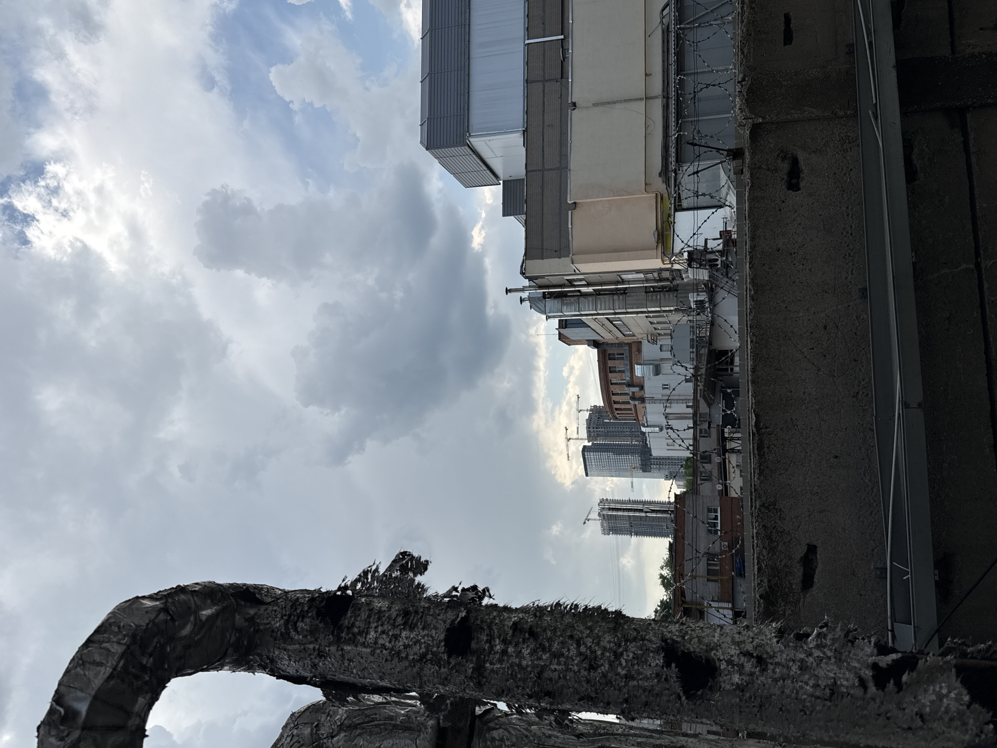 The image shows a view upwards towards the sky in Khimki, Russia. The foreground consists of a dark, damaged, possibly burned structure. Behind it are buildings, including some with barbed wire along the roofline. Further in the distance, several high-rise buildings are under construction, with cranes visible. The sky is cloudy.