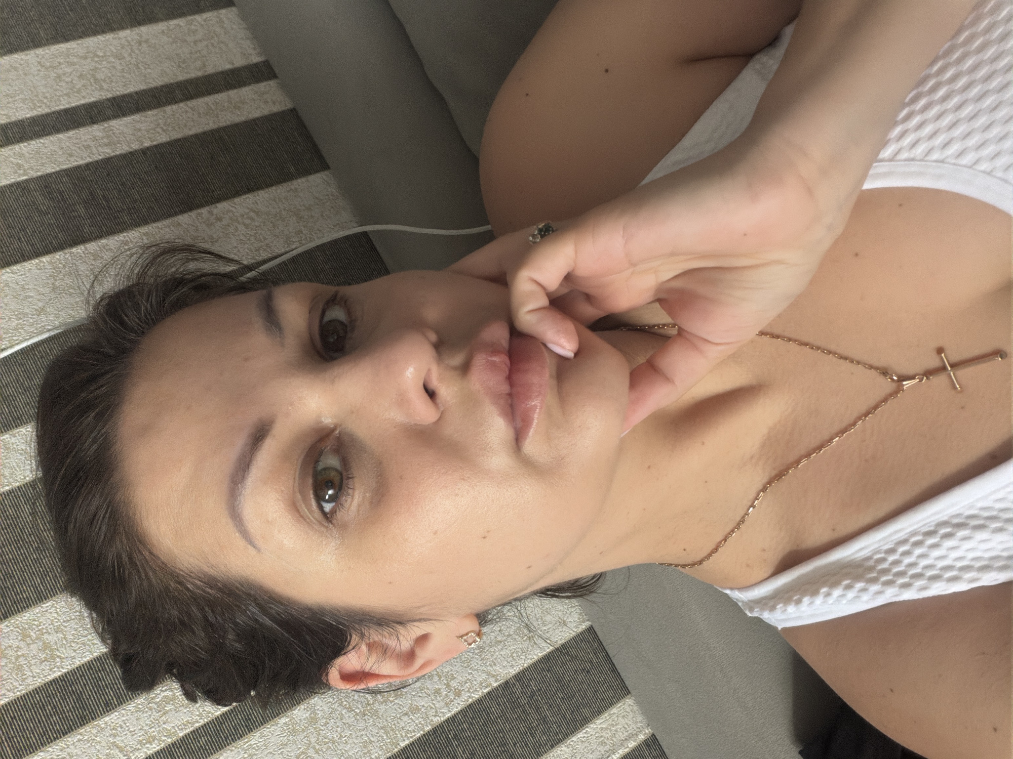 A selfie is taken from a close-up, high-angle perspective of a woman with dark hair pulled back. She is wearing a white tank top and a gold necklace with a cross pendant. Her left hand is resting on her chin, and she is wearing a ring on her right index finger and an earring in her left ear. She is lying on a surface that appears to be a couch or bed with a striped fabric headboard in the background. The lighting suggests an indoor setting.