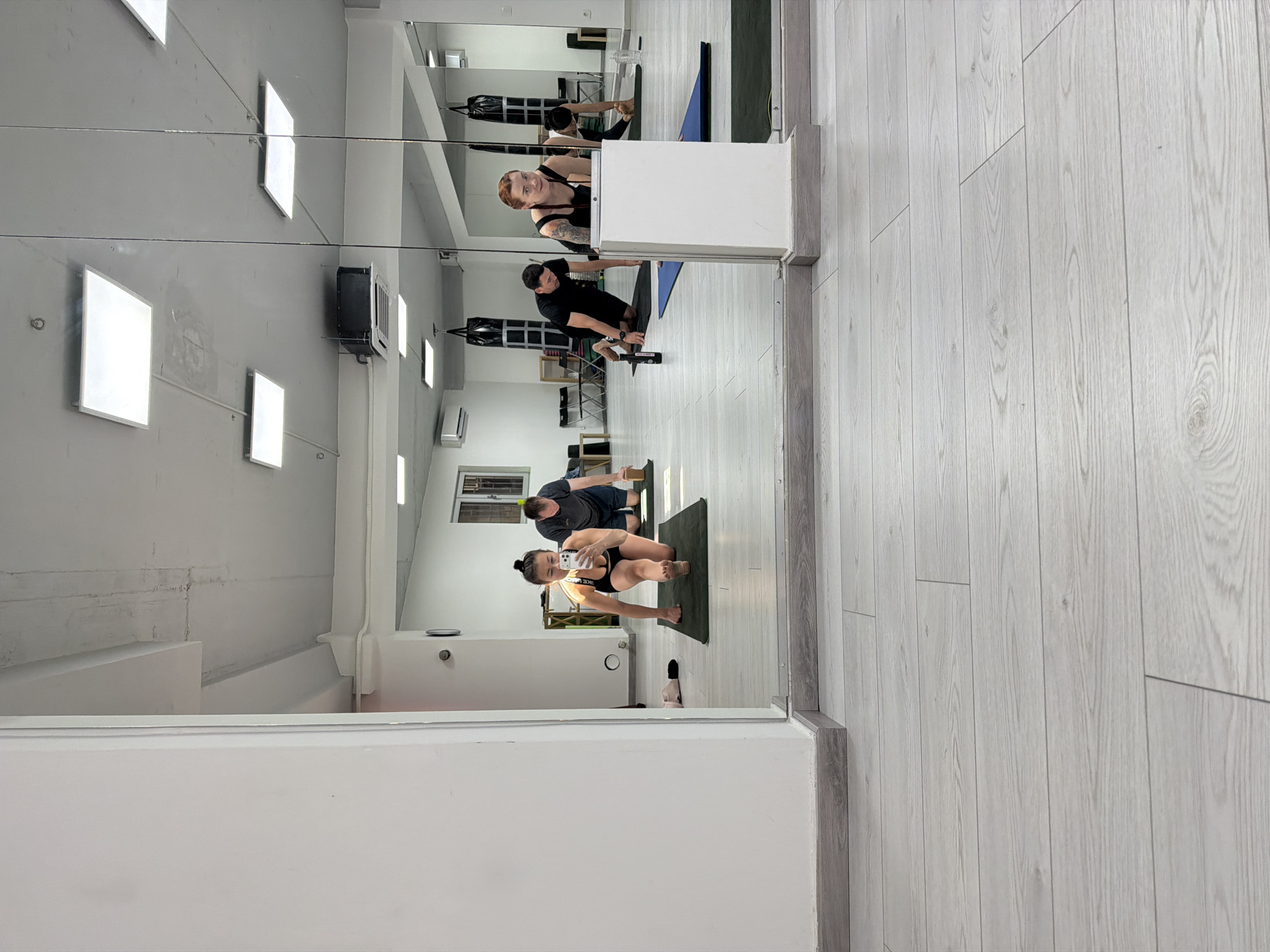 The image captures the reflection in a mirror within a fitness studio in Budva, Montenegro. A woman is taking a selfie with her phone, situated at the forefront of the reflection. Behind her, a man is visible on a mat, seemingly engaged in exercise. Further in the mirror's reflection, there are two more individuals, a woman and a man. The studio appears well-lit and equipped with workout equipment. The floor is covered with a light grey wooden plank design. The overall scene conveys an active and health-conscious environment.