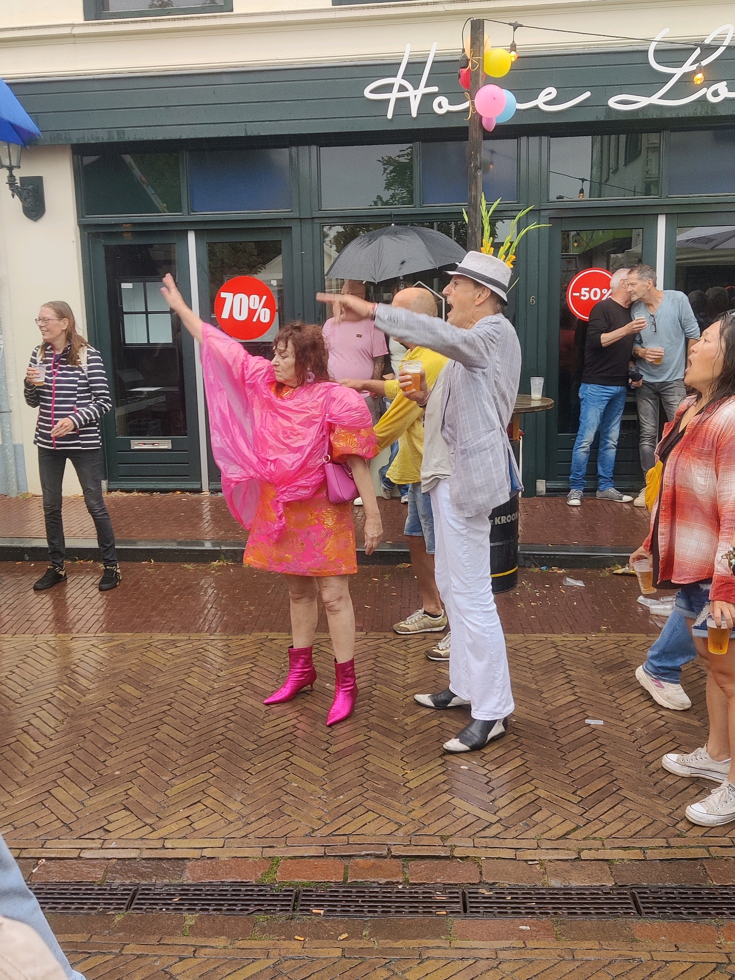 A group of people are gathered outside of a building in Winterswijk, Netherlands, on a wet brick sidewalk. The building has a dark green facade with large windows and the word "Horeca" and "Lo" visible in white script lettering above the entrance. Two red circular signs with percentages, "70%" and "-50%", are displayed on the windows. A woman in a bright pink poncho and orange dress with matching pink boots is dancing and gesturing with her hands. She is holding a pink purse. To her left, a woman in a striped jacket and black pants is holding a drink. A man in a hat and light-colored suit is pointing to the left and holding a drink. Behind him, several other individuals are visible, some holding drinks, and one is under a black umbrella. The ground is wet, indicating recent rain. Festive balloons are attached to a pole near the entrance.