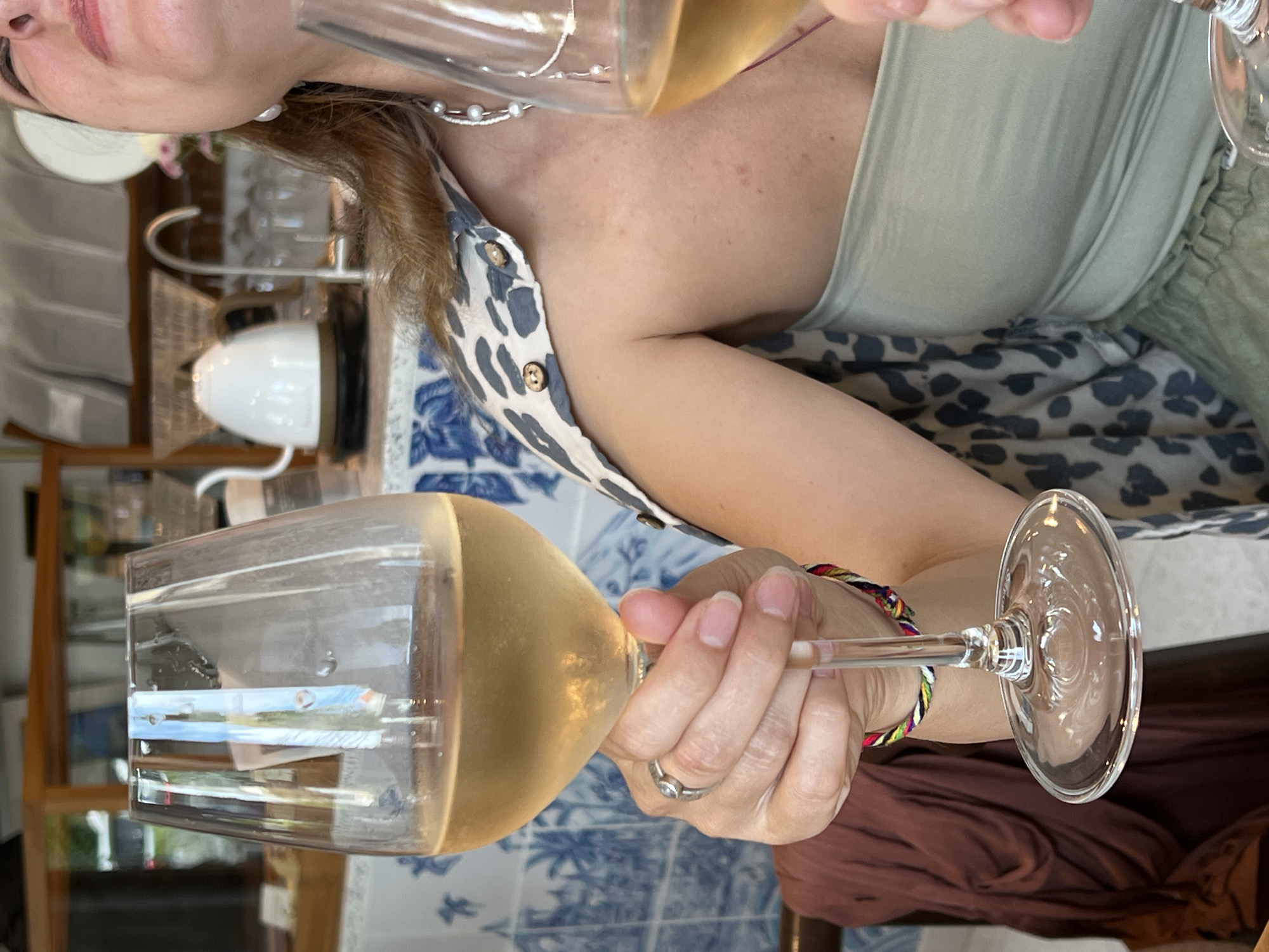 The scene depicts a person holding a wine glass filled with a light-colored liquid, likely wine, indoors. The person is wearing a top with a leopard print and a light green tank top beneath it. Their hand holding the glass has a ring on the ring finger and a colorful braided bracelet. The background includes a blue and white patterned surface and a glimpse of a room with shelves and various objects. The scene is indoors, and based on the available information, it is not possible to confirm the location in the Ubud District, Indonesia.