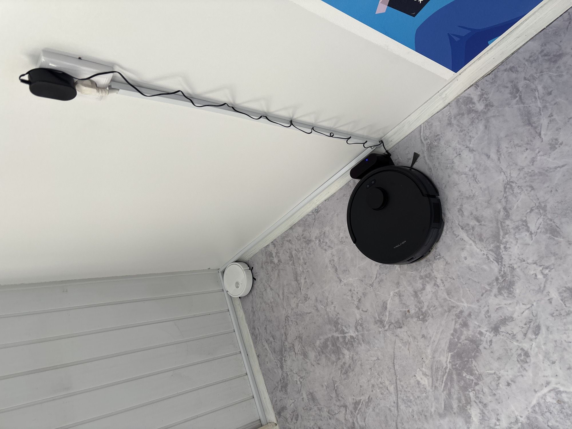 In a corner of a room with white, vertically paneled walls and a grey marble-patterned floor, a black robot vacuum cleaner is parked on its charging base. The charging base is plugged into a white electrical outlet on the wall, which also has a black adapter plugged into it. A black cord snakes down the wall from the adapter and connects to the charging base. Further back in the corner, near the wall, sits a white robot vacuum cleaner. A small blue light is visible on the black robot's charging base, indicating it is charging. To the right of the charging area, a portion of a colorful graphic poster is visible on the wall. The image appears to be taken indoors, in a domestic setting, possibly a home or apartment. The scene does not contain any information that would allow for its specific location in Vileyka, Belarus.