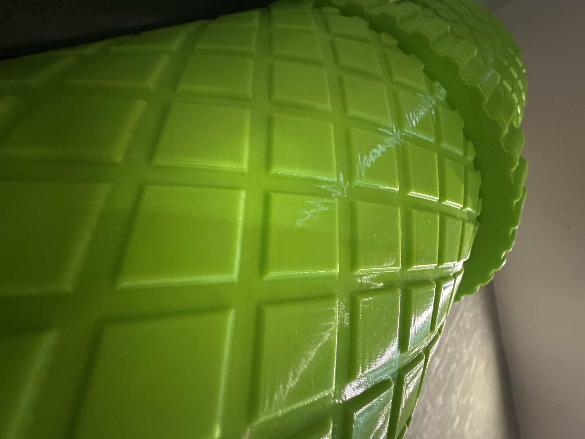 The image is a close-up, high-angle shot of a bright green plastic object with a textured, grid-like pattern. The object appears to be a container, possibly a bucket or pot, with a scalloped edge visible at the top. The lighting creates highlights and shadows across the surface, emphasizing the raised square and rectangular sections of the texture. There are some faint scuff marks or imperfections visible on the surface of the plastic. The background is dark at the top and transitions to a lighter, neutral color on the right side, suggesting an indoor environment. No people or any indication of a specific location in Moscow, Russia are present in the image.