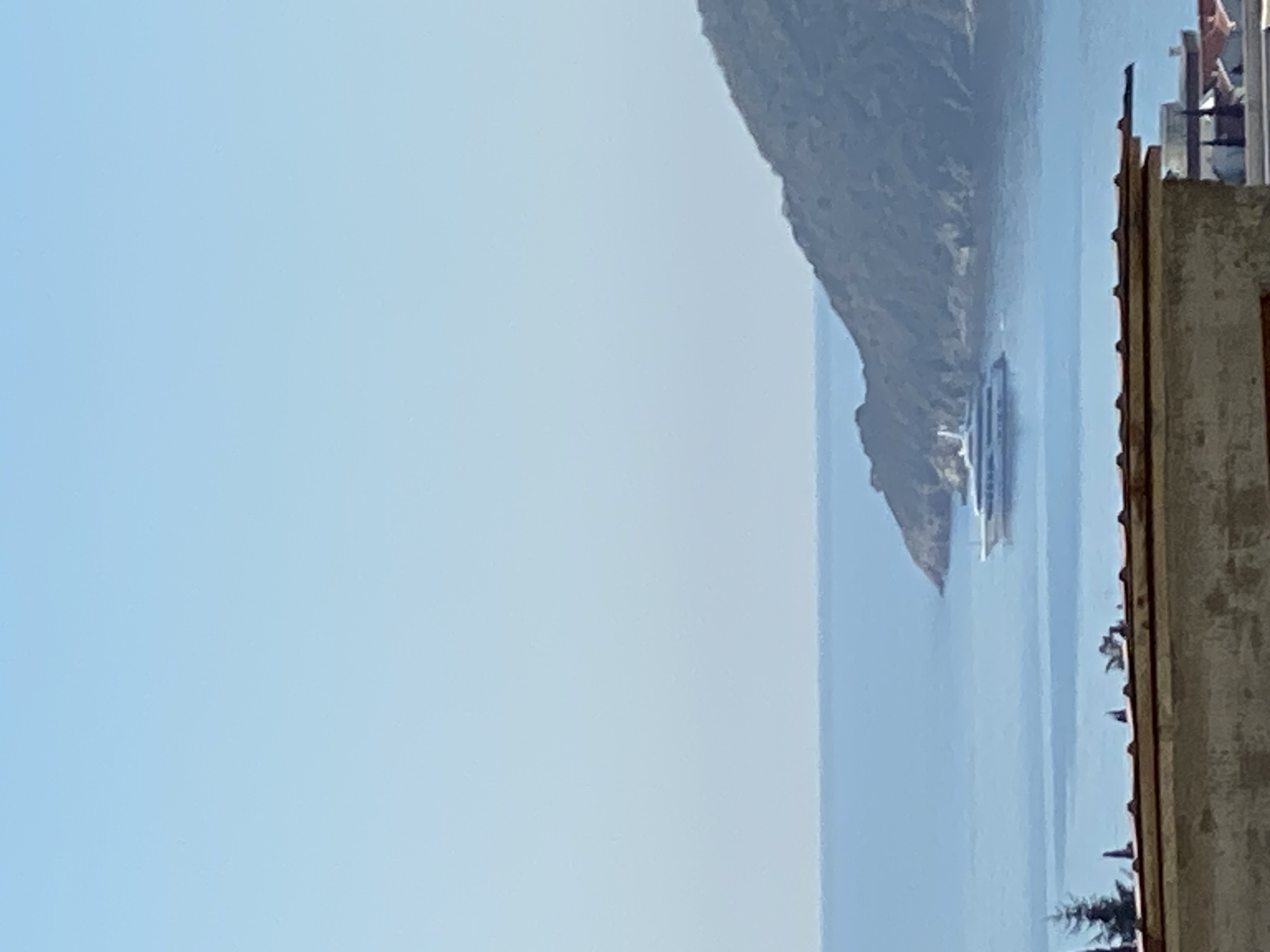 The image shows a view of the Bay of Kotor in Budva, Montenegro. The composition includes a mountainous shoreline, a ship on the water, a clear sky, and the edge of a building.