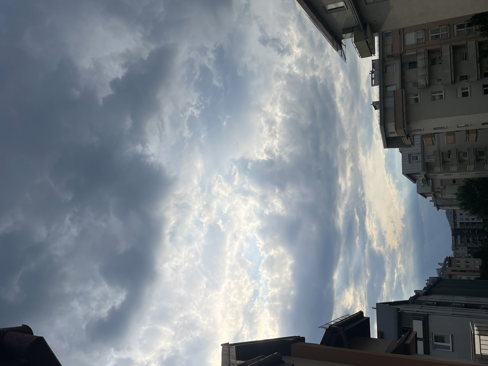 The image shows a view of apartment buildings against a cloudy sky. The buildings are of varying heights and architectural styles, with balconies visible on some of them. The sky is filled with a mix of dark and light clouds, suggesting an overcast or impending stormy weather. The composition is from a slightly elevated perspective, looking upwards towards the sky, with the tops of the buildings framing the lower part of the image. There are no people or animals visible in the scene. The environment appears to be urban, with multiple residential structures.