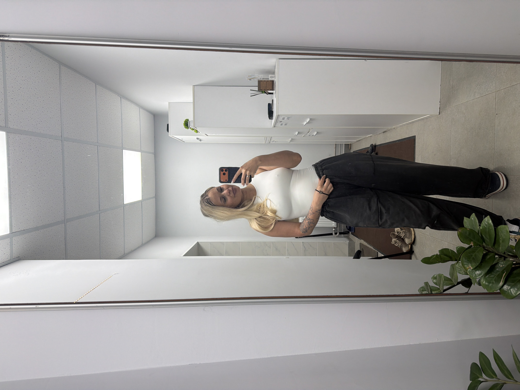 A young, fair-skinned woman with blonde hair is standing in front of a large mirror and taking a selfie with her smartphone. She is wearing a white tank top and loose black pants. Her left arm has tattoos. The reflection in the mirror shows a room with white walls, a tiled ceiling, and a light-colored tile floor. Along the wall are white storage cabinets. A dark brown doormat is visible on the floor. There is a green plant in the lower right corner of the image. The location is identified as Sarapul, Russia.