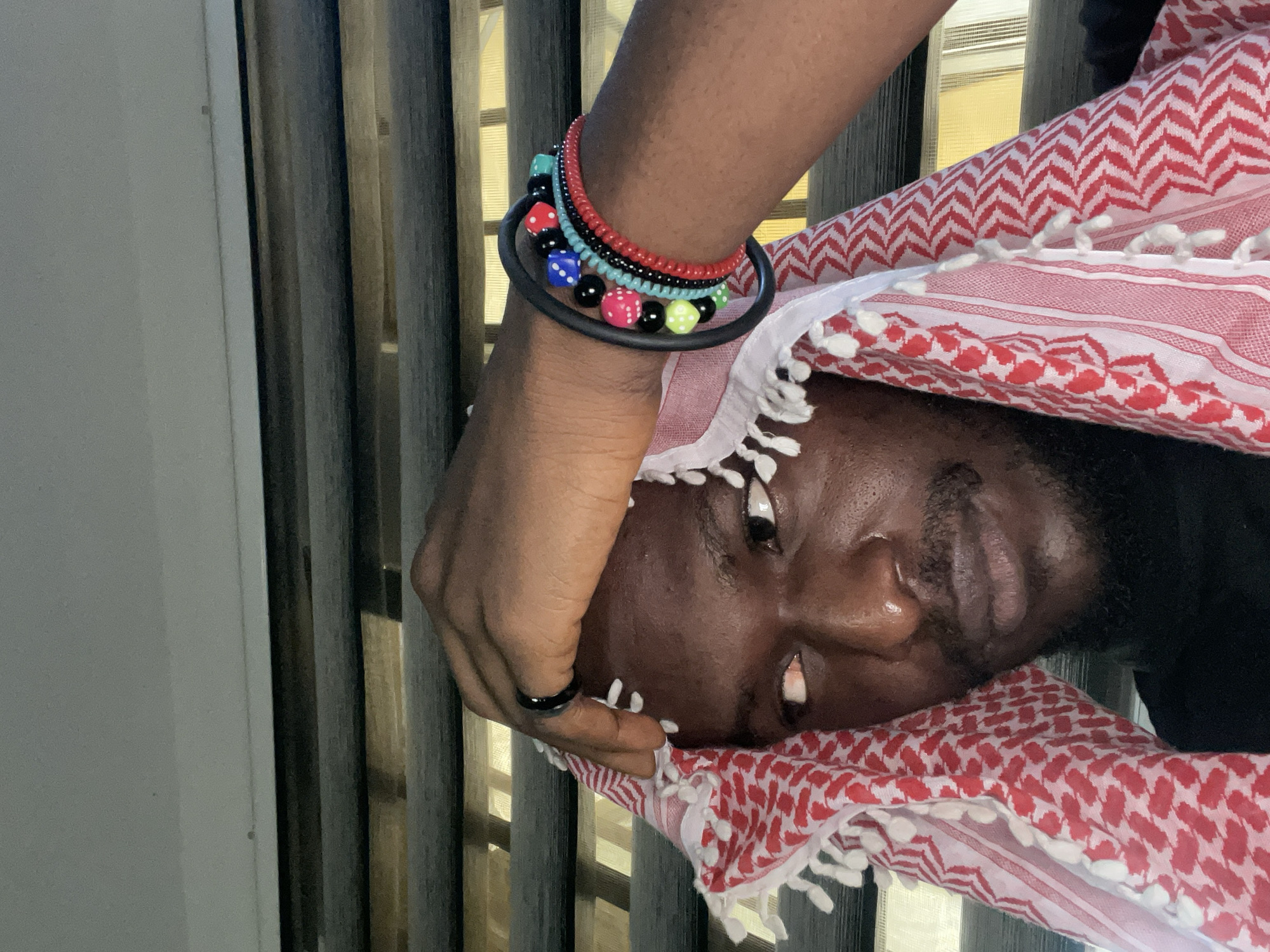 A close-up shot shows a Black man wearing a red and white keffiyeh. He has a black ring on his left hand, which is resting on his forehead. He is also wearing several beaded bracelets on his right wrist, including some shaped like dice. The man is looking to his right with his eyes slightly narrowed. The background appears to be a window with horizontal blinds. The image quality is fair with some noise. The overall impression is a portrait, likely taken indoors.
