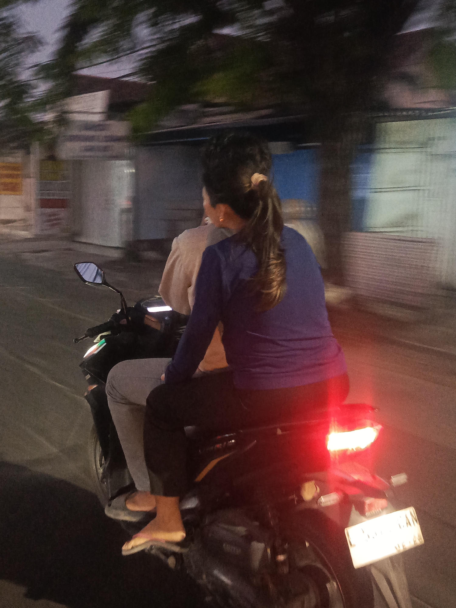 The image captures a motion-blurred view of two people riding a motorcycle on a street at dusk, likely in Surabaya, Indonesia. The rider in front is wearing a light-colored long-sleeved shirt, while the passenger behind wears a dark blue long-sleeved shirt. The passenger's hair is pulled back in a ponytail. The motorcycle has a visible taillight illuminated in red. The background consists of blurred buildings and trees, suggesting an urban or semi-urban environment. The license plate of the motorcycle is partially visible, with characters that appear to be in the Indonesian format, further supporting the location. The overall lighting suggests it is either early morning or evening.