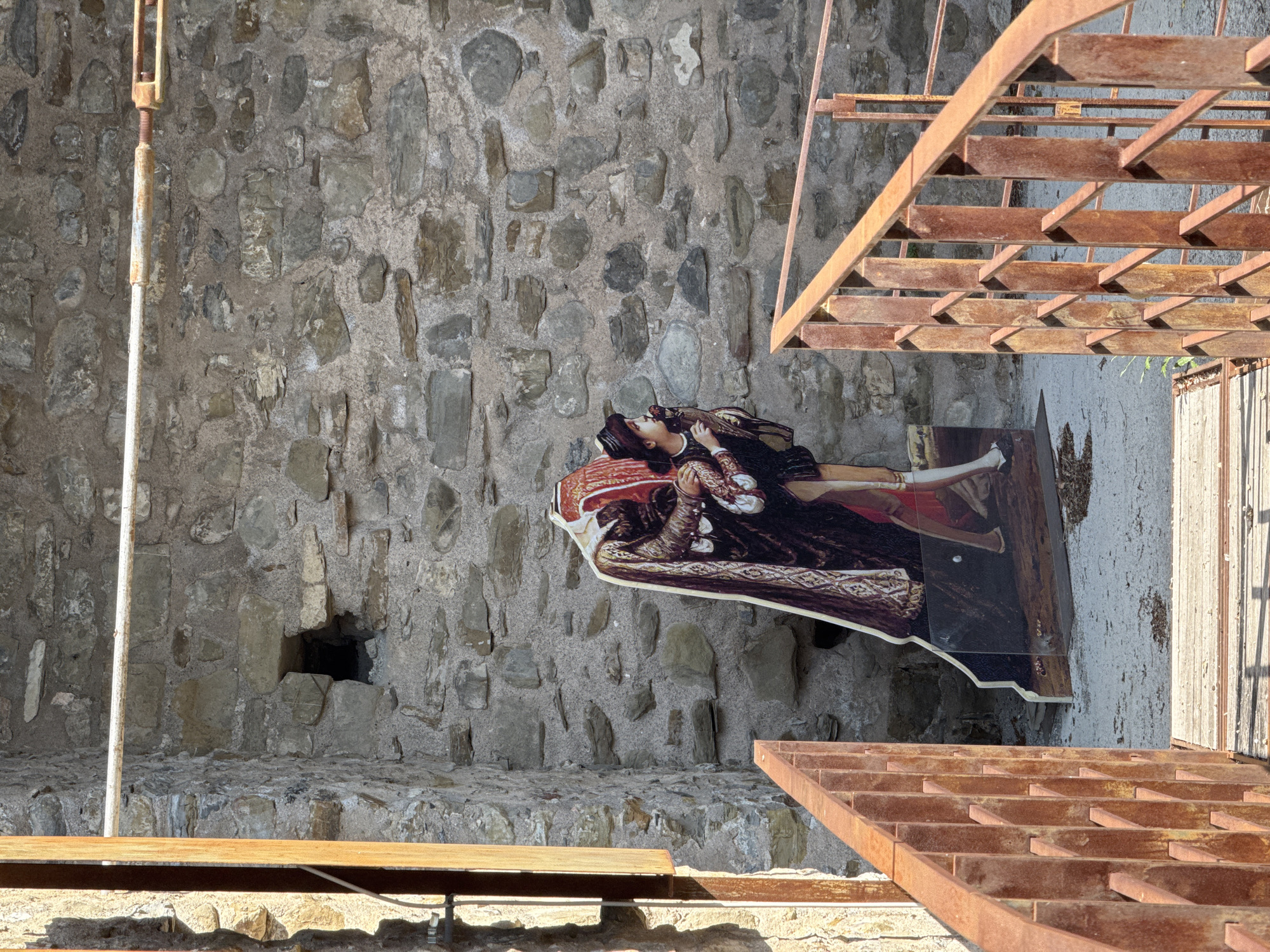 A cutout figure depicting a man in historical attire stands in front of a stone wall, partially obscured by a rusty metal railing. The figure's head is cropped out of the frame, but the elaborate cloak and the lower half of the man's body, including his legs and feet in stockings and shoes, are visible. He is shown from the waist down, with his right hand touching a bird perched on his left hand. The background is a rough stone wall with an opening to a dark space. A narrow wooden walkway with a rusty metal railing leads towards the figure. The scene is located in Dolceacqua, Italy, near the Doria Castle.