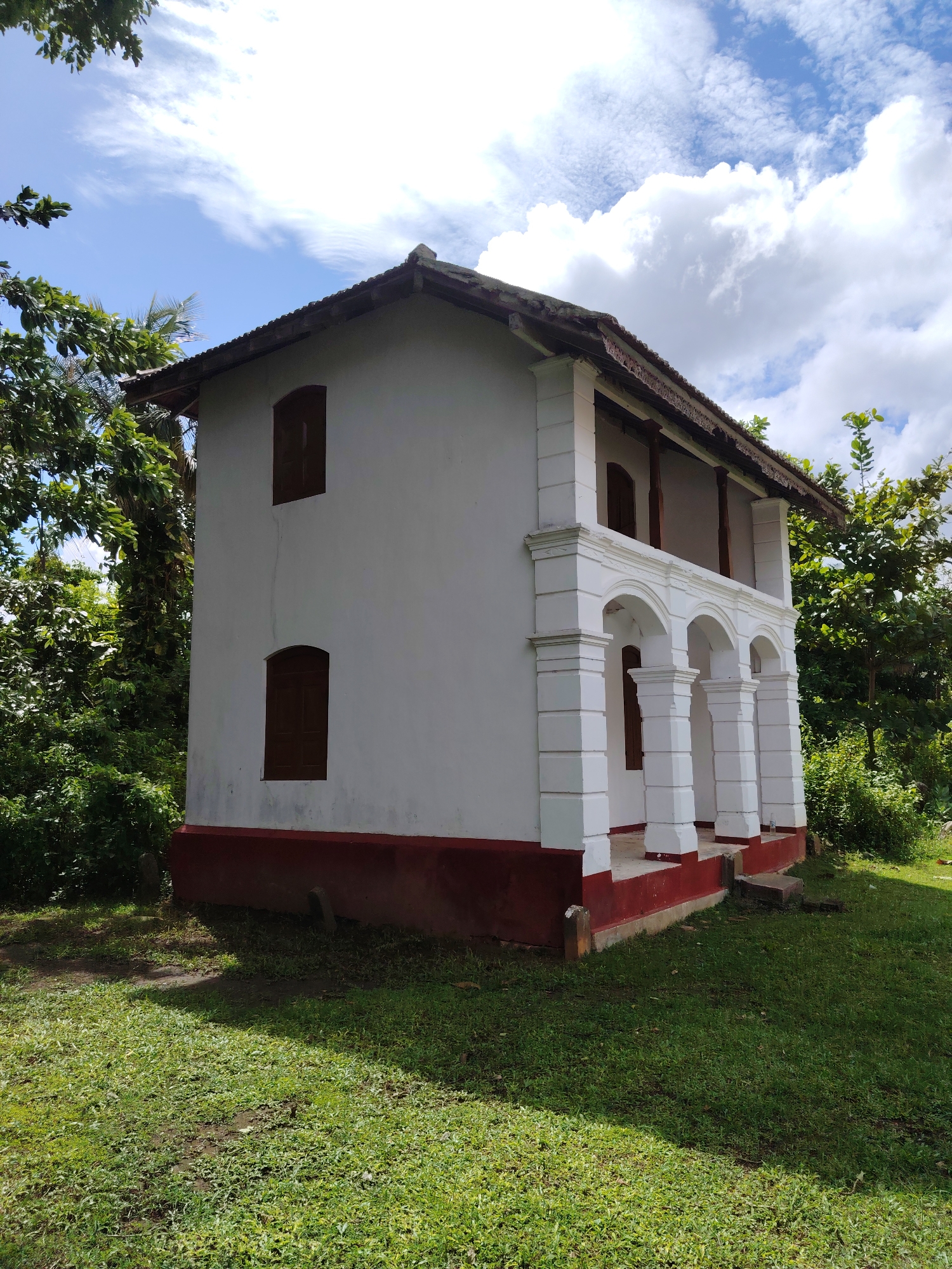 A two-story, white-walled building with a red base is the central subject, set amidst lush green grass and dense foliage under a partly cloudy blue sky. The building exhibits a traditional or colonial architectural style.
The front of the building features a porch area with multiple white, arched columns supporting an upper-level balcony. Three arched columns are clearly visible on the ground floor, leading to a covered entrance or veranda. The upper balcony has a simple railing supported by dark wooden posts. On the left side of the building, two dark brown wooden windows with closed shutters are visible, one on each floor. Additional dark wooden elements, likely doors or windows, are visible behind the arched columns on the porch. The roof is tiled, appears weathered, and has a significant overhang.
The building is surrounded by vibrant green grass in the foreground, with some patches of darker soil. Tall, leafy green trees and thick bushes frame the structure, particularly on the left and rear, suggesting a verdant, possibly tropical environment. The sky is bright blue with scattered white cumulus clouds, indicating fair weather. The time of day is daytime, evidenced by the bright illumination and shadows cast on the grass. No people or visible text are present in the image. The scene conveys a sense of peaceful solitude and historical character.