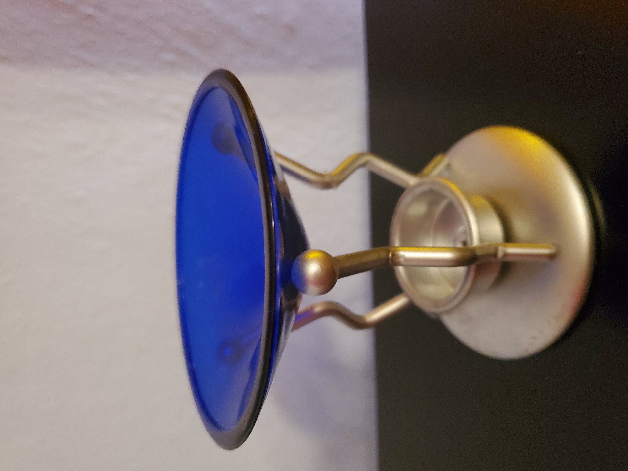An oil burner with a blue glass dish and a silver metal stand is placed on a dark, reflective surface. The background is a plain, light-colored wall. The oil burner is the sole object in the frame. There are no people, animals, or visible actions taking place. The scene appears to be indoors, in a domestic or commercial setting, with no discernible connection to the city of Konstantinovka, Moldova.