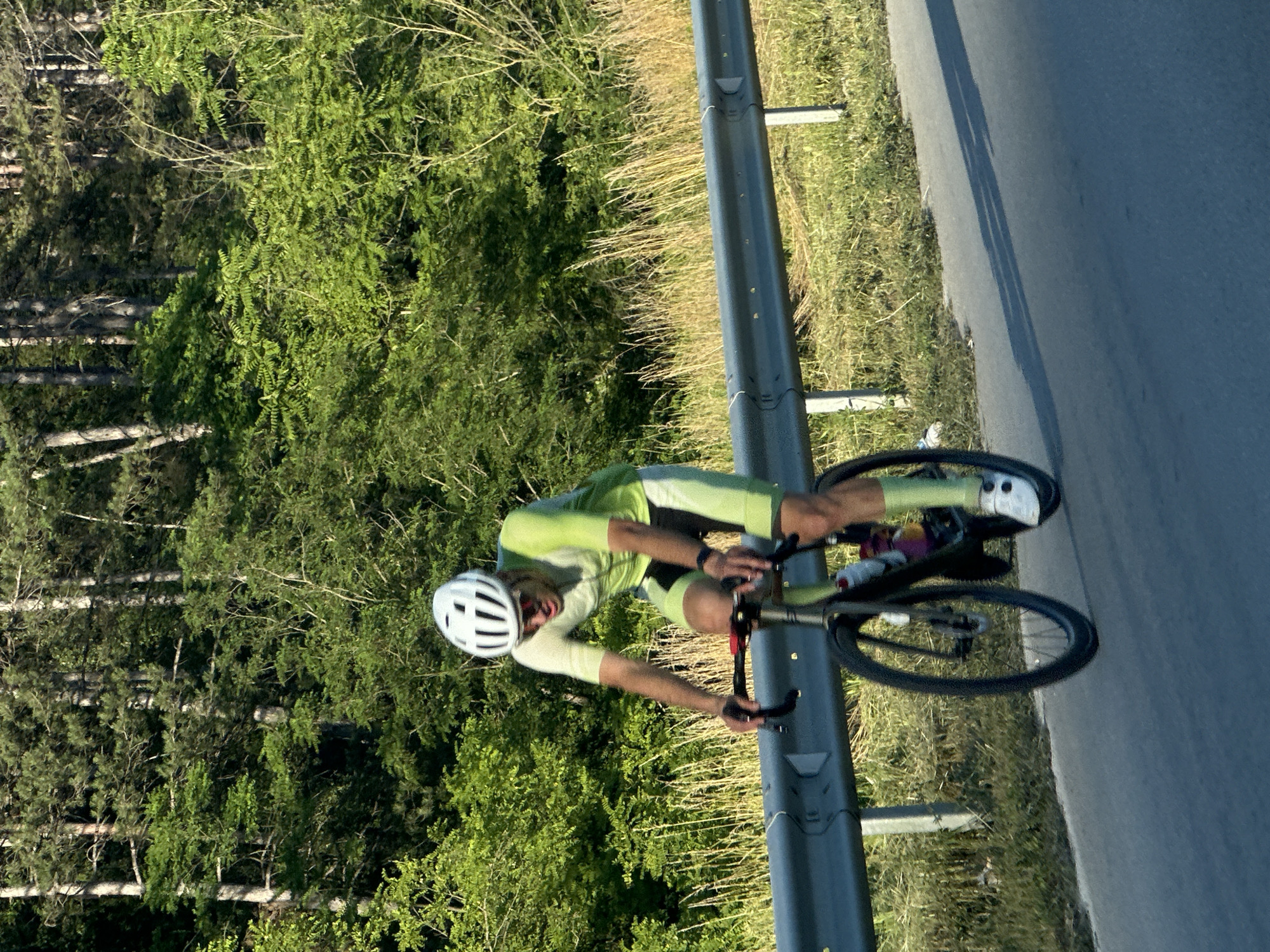 A person is riding a bicycle on a road bordered by a metal guardrail and lush green trees. The cyclist is wearing a white helmet and a light green cycling suit, with bright yellow accents on the shorts. The rider's left leg is extended, pushing down on the pedal, while the right leg is bent, completing the circular motion of cycling. Both hands are on the handlebars.
The scene appears to be set during the daytime, likely late morning or early afternoon, given the bright sunlight casting shadows on the road. The weather seems fair and warm, as indicated by the vibrant greenery and the cyclist's attire. The background consists of dense trees with some dry grass and weeds growing along the roadside. The road itself is asphalt and appears to be a paved route, possibly in a rural or semi-rural area given the natural surroundings.
The overall impression is one of athletic activity and a serene, natural environment. The focus is on the cyclist's movement and the dynamic action of riding. There is no visible text in the image. The setting, with the guardrail and trees alongside a road, suggests a cycling route in a scenic location, possibly in Bulgaria, near Prisovo, as per the location context.