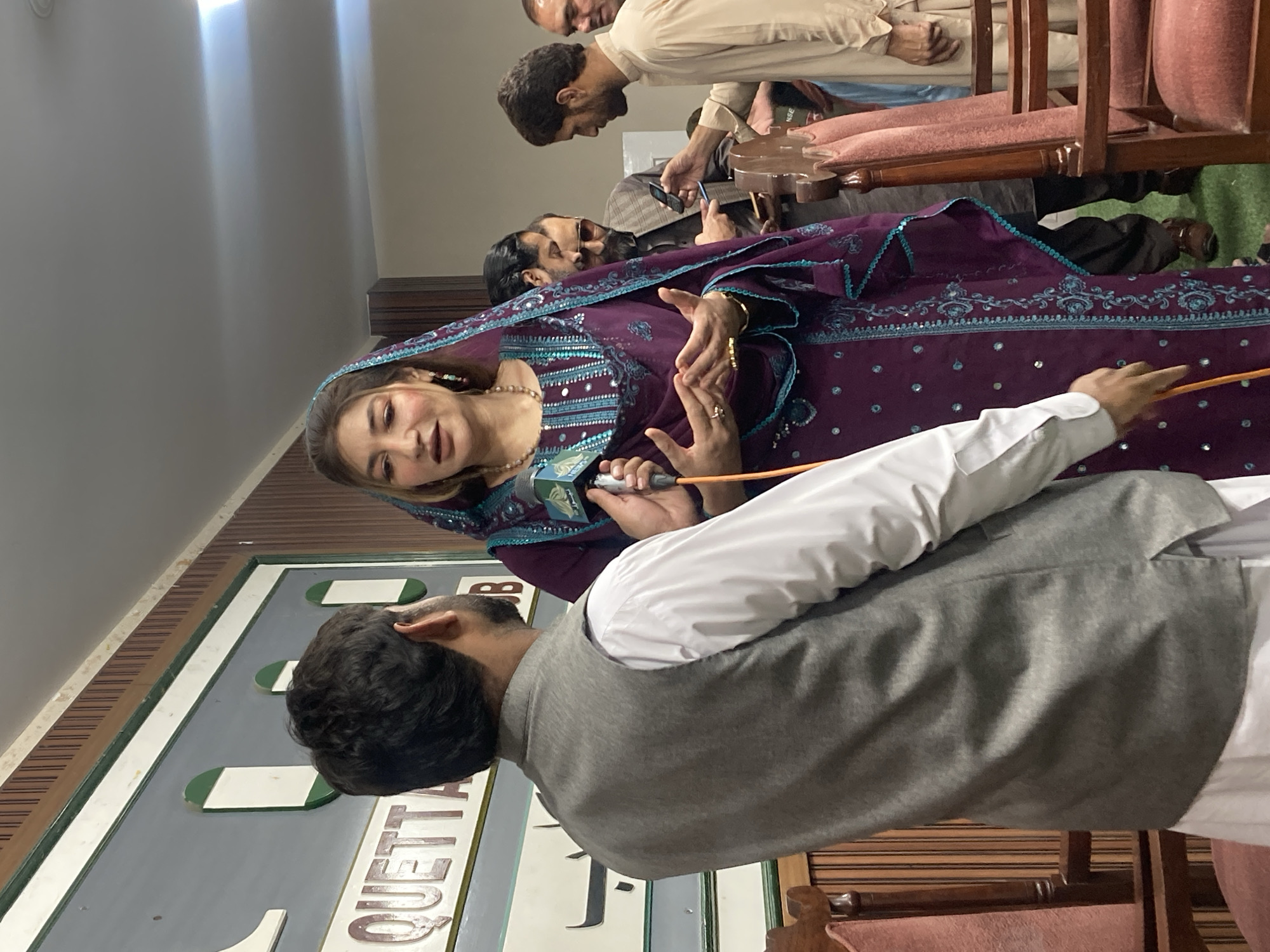 Inside a room in Quetta City Tehsil, Pakistan, a woman in a purple, intricately embroidered garment and headscarf is speaking into a microphone held by a man in a white shirt and gray vest. Other individuals are visible in the background, some seated, with one using a mobile phone. The background includes a sign partially visible that says "Quetta". The room features wooden paneling and what appears to be office or institutional seating.