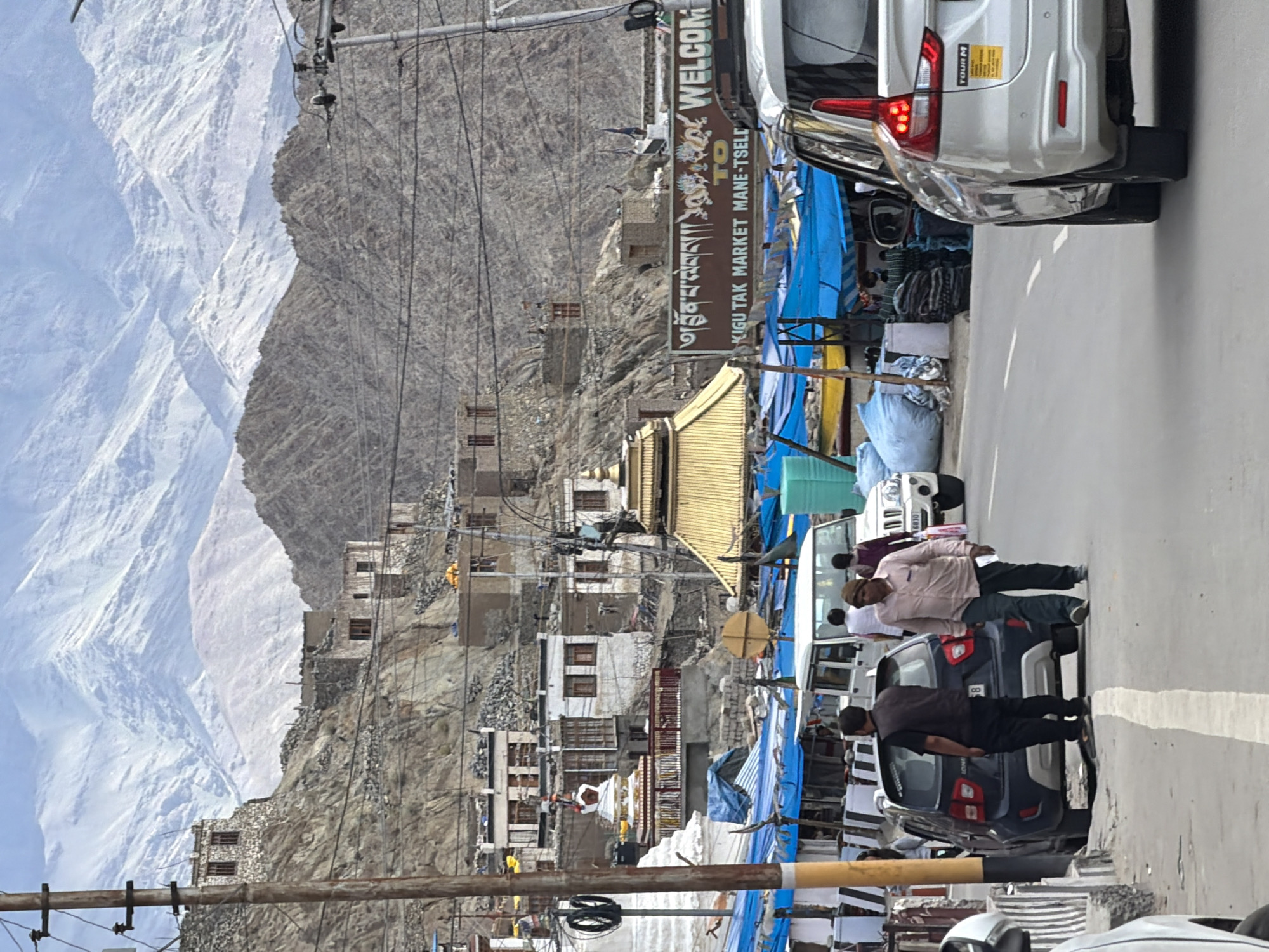 The image depicts a street scene in the city of Leh, India, with a backdrop of majestic snow-capped mountains. In the foreground, several cars are visible, including a white MPV and a dark blue hatchback. Two men are walking on the side of the road, with one of them in a light-colored shirt and dark pants, and the other in a dark vest and pants. Behind them, a bustling market area is set up with blue tarps covering stalls, displaying various goods. Buildings are built on the rocky slopes of the mountains in the midground. A prominent sign with Tibetan script and the English text "WELCOME TO KIGU TAK MARKET MANE-TSEL" is affixed to a building. The overall atmosphere suggests a vibrant street scene in a mountainous region.