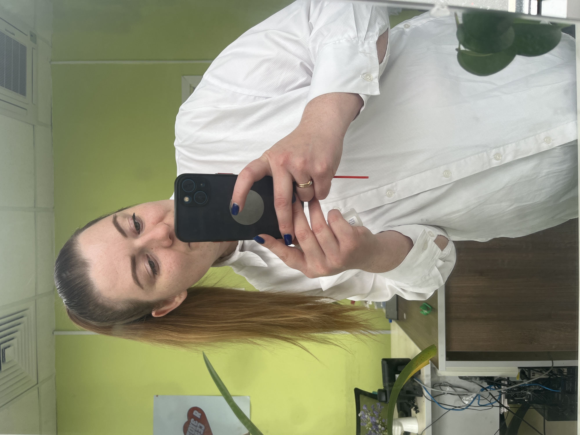 The image captures a mirror selfie taken by a woman in what appears to be an office or workspace located in Klin, Russia. The woman, with reddish-brown hair pulled back into a ponytail, looks directly into the smartphone camera held in her left hand. She has light skin, visible freckles on her nose and cheeks, and a neutral to slightly smiling expression. Her left hand, adorned with blue nail polish, holds a black smartphone (resembling an iPhone model with three prominent camera lenses on the back). A gold-colored band ring is on her left ring finger. She wears a white, long-sleeved collared shirt with a subtle vertical stripe pattern. Her right hand holds a small white bottle, and a thin red stylus or similar object is visible between her fingers.
The background, a reflection in the mirror, features light green walls. In the upper left corner of the reflection, a white air conditioning vent with a grille is mounted. Toward the upper right, a plant with several broad green leaves is reflected. The lower right portion of the background shows a wooden desk surface, beneath which are visible electronic equipment and a tangle of blue and black wires. Another plant with a single long, pointed green leaf extends into the lower left foreground of the reflection. A white sign or board with a partially obscured red logo, possibly "TOYS R US" or similar text, is visible on the lower left wall. The indoor lighting appears bright, suggesting daytime.