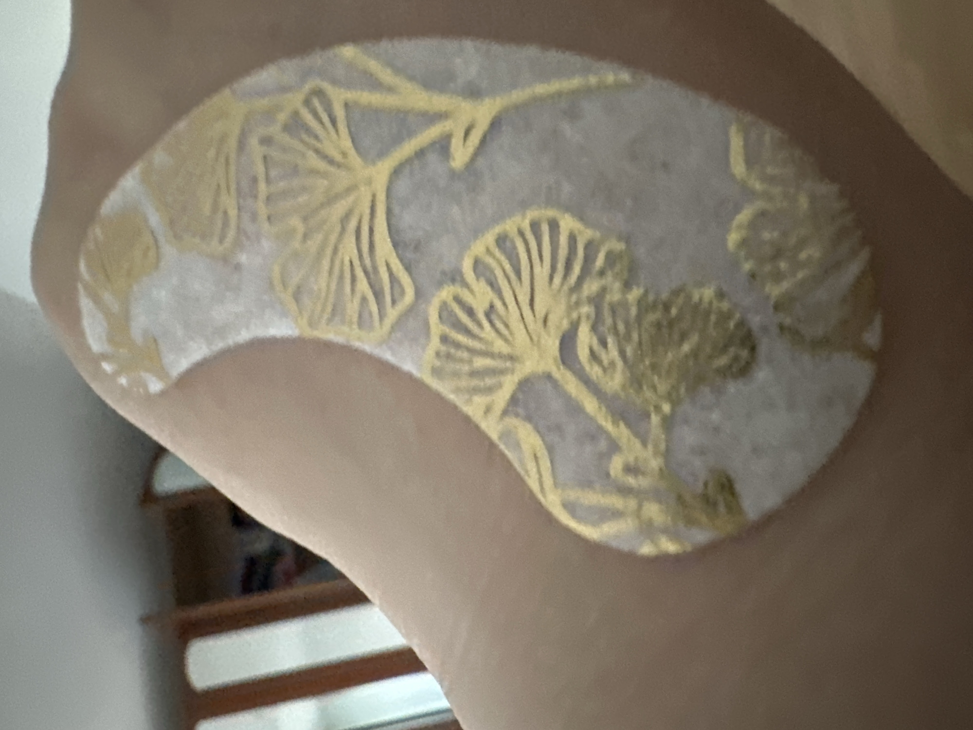 An object, likely a bandage or decorative patch, with a pattern resembling ginkgo leaves in gold is adhered to a person's skin. The patch is oval-shaped and appears to be positioned on an arm or shoulder. In the blurred background, wooden furniture, possibly a cabinet or chair, and a ceiling are visible, suggesting an indoor domestic setting. The location is Krefeld, Germany.