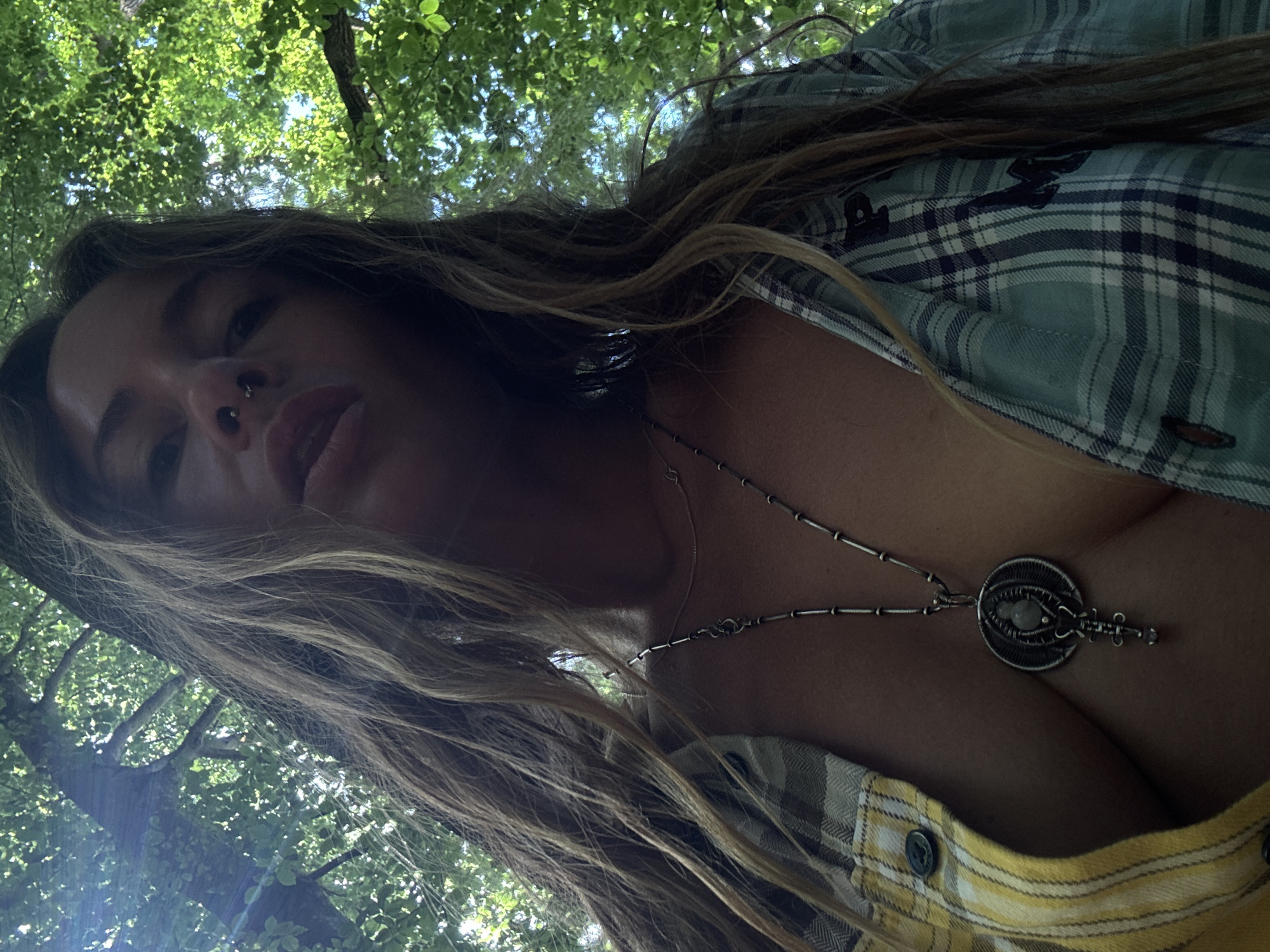 A low-angle, medium shot captures a woman from the chest up, looking directly at the camera. She has long, wavy blonde hair that cascades over her shoulders and chest, partially obscuring her face and clothing. Her face is tilted downwards, with her eyes looking up towards the viewer. She has a septum piercing and her lips are parted. She is wearing an open plaid shirt, revealing her chest and a necklace with a circular pendant. The background is dominated by the lush green foliage of trees, with dappled sunlight filtering through the leaves, indicating an outdoor setting, likely in a park or wooded area. The lighting is natural and bright, suggesting it is daytime.
