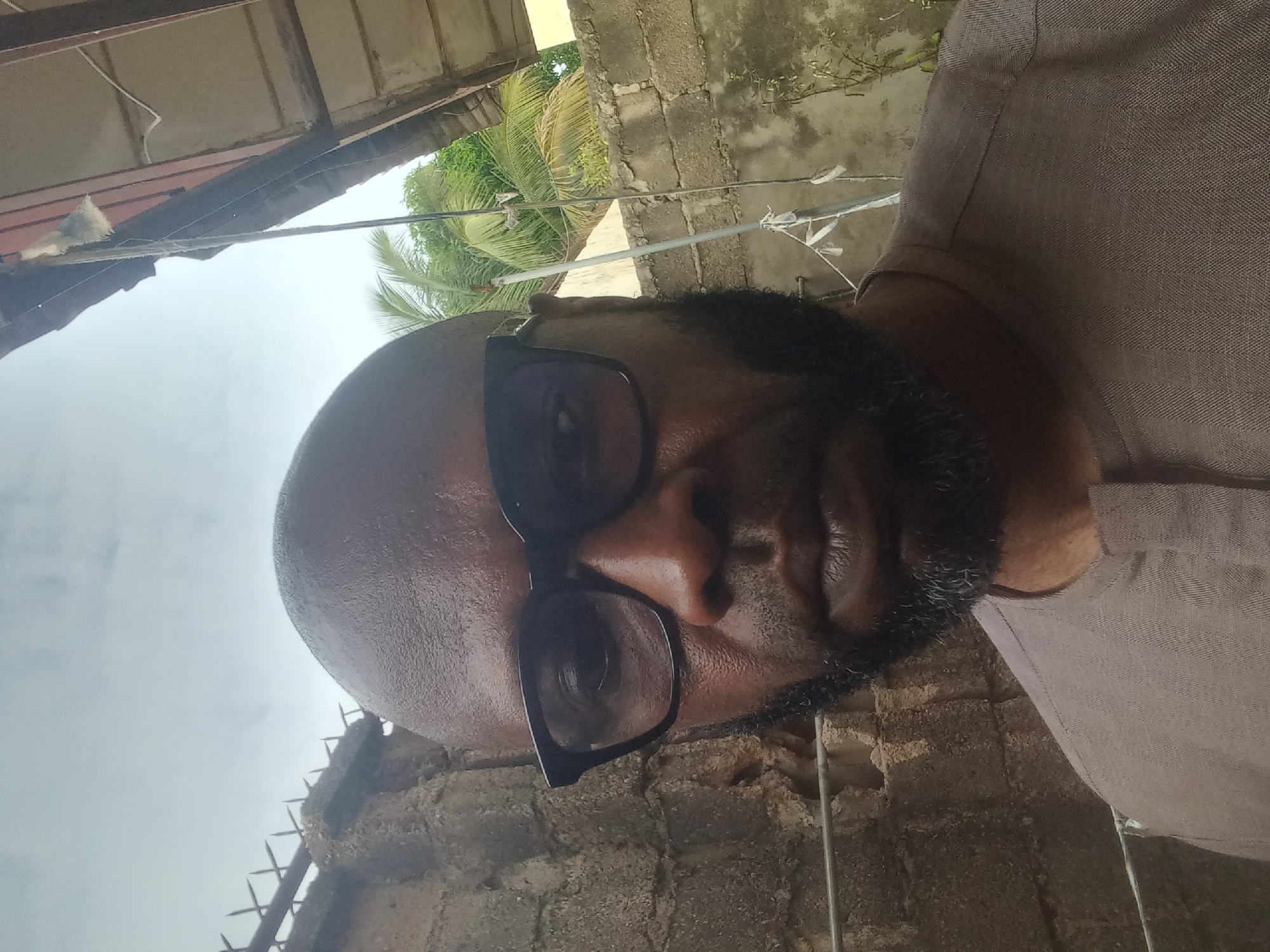 The image, captured from a slightly low angle and presented rotated 90 degrees counter-clockwise, is a close-up portrait of a person from the chest up, looking directly at the viewer. The individual, appearing male, has a bald head and a dark beard, and wears dark-rimmed glasses. He is dressed in a light brown or beige collared shirt with a subtle textured pattern. His skin shows a slight sheen, possibly from warmth or lighting.
The background indicates an outdoor setting under an overcast sky during daytime. Above the person's head, visible elements include an awning or roof structure made of corrugated material and wood, with utility wires strung across. To the upper right (left in the rotated image), lush green palm foliage is present. Below the person and extending to the lower left (bottom in the rotated image), a concrete block wall forms part of the environment. A barbed wire or spiked fence structure is visible atop this wall. The setting appears to be a functional, possibly residential or semi-urban area, consistent with the Barden Yana, Nigeria context, featuring common building materials and tropical greenery. No text is visible.