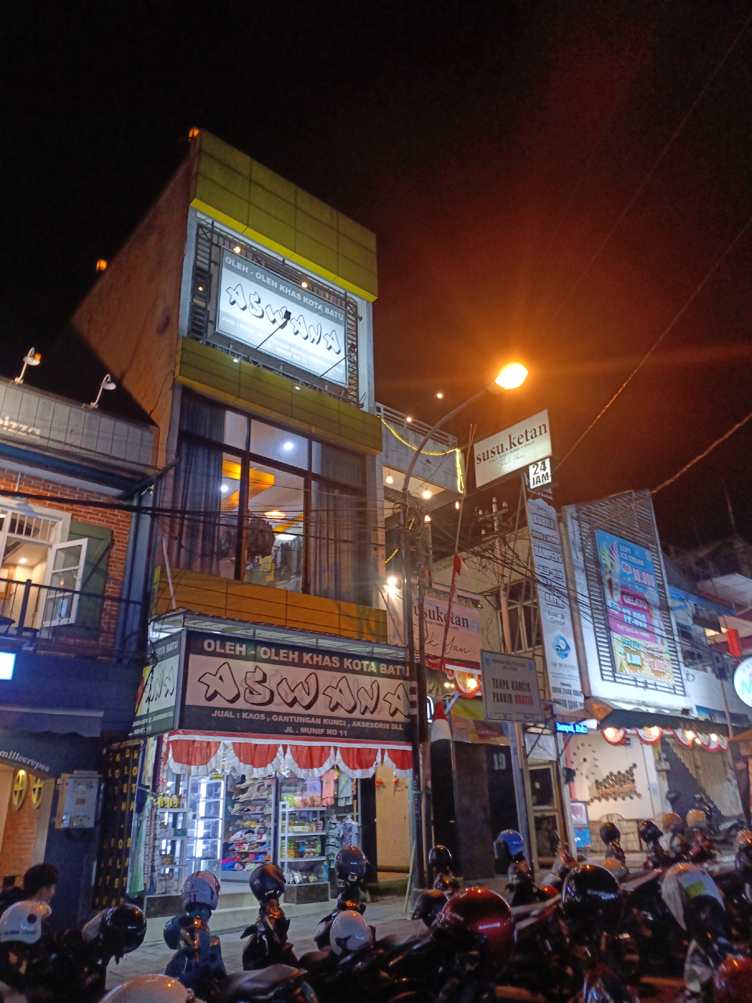 The image depicts a street scene at night in the city of Batu, Indonesia. Several buildings line the street, illuminated by neon signs and streetlights. On the left, a multi-story building displays a prominent sign that reads "ASWANA OLEH-OLEH KHAS KOTA BATU," indicating it's a souvenir shop specializing in goods from Batu. Below this sign, the shop's storefront is visible, filled with shelves of products. In front of the shop and extending down the street, numerous motorcycles are parked, many with helmets placed on them. Several individuals are visible, including a person standing near a motorcycle on the far left, and others partially obscured in the background. Further down the street, another building has a sign for "SUSU KETAN" (sticky rice milk) with "24 JAM" (24 hours) indicating it's a 24-hour establishment. The overall atmosphere is one of a bustling commercial area at night, with a focus on local businesses and transportation.