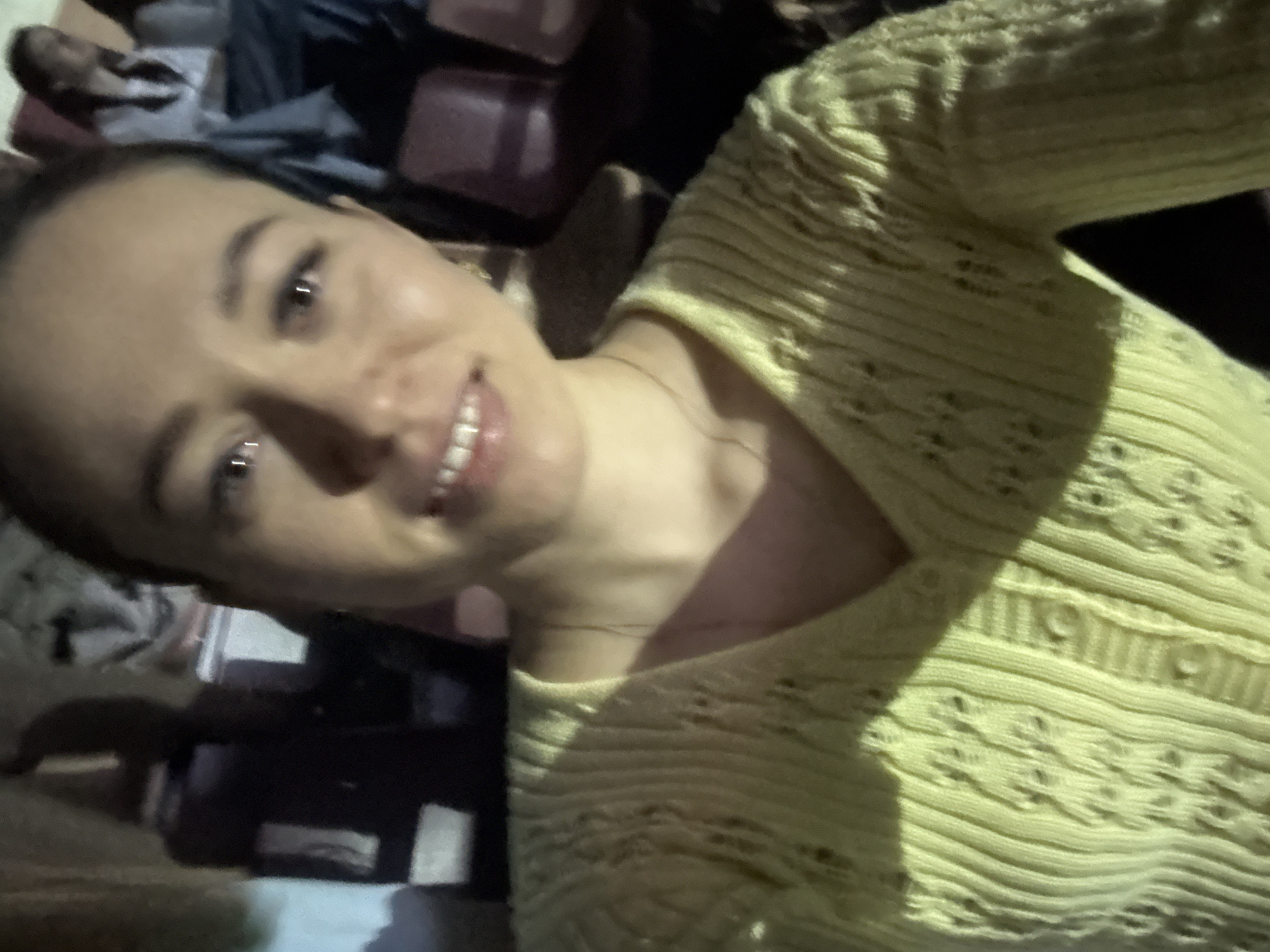 A person with light skin and dark hair is smiling and looking at the camera. They are wearing a yellow, textured v-neck top. The background appears to be a cluttered space with luggage or bags visible. There is insufficient information to determine the location or confirm it is in Perm, Russia.