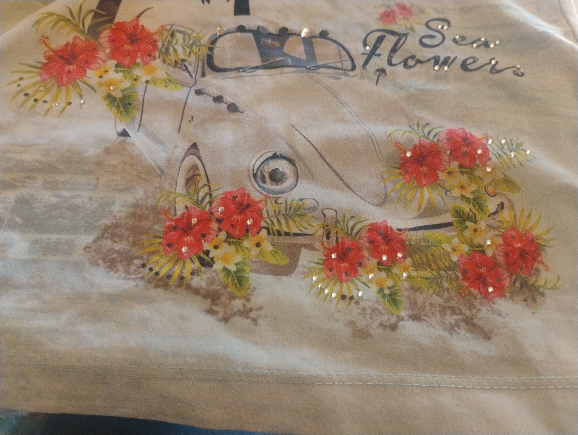 The image shows a close-up of a light-colored t-shirt with a graphic print. The graphic features a vintage car adorned with red hibiscus flowers and greenery. The words "Sea Flowers" are printed in a stylized font above the car, with small rhinestones accenting the letters. The t-shirt itself has a textured appearance and a visible seam running horizontally across the bottom. The lighting is soft, and the overall impression is of a casual, summery garment. The location in Rumyantsevo, Bulgaria, is not discernible from the image itself.
