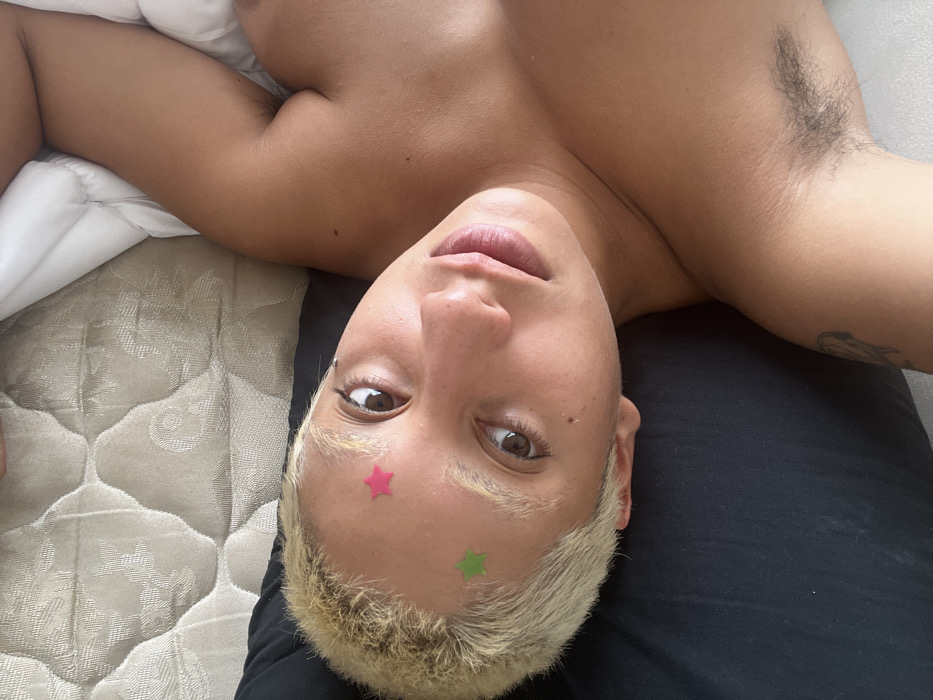 A close-up, high-angle shot depicts an individual with fair skin and short, bleached blonde hair, lying down and gazing directly into the camera. A pink star-shaped sticker is affixed to the upper forehead, with a green star-shaped sticker positioned below and slightly left. The person's dark eyes are focused, and lips are slightly parted. The individual is bare-chested; the right shoulder is visible, and dark axillary hair is evident in the left armpit. A faint tattoo is present on the upper left arm. The background features a light-colored quilted mattress pattern in the upper left, a dark fabric (likely a pillow) beneath the head, and white bedding along the right side of the frame. The scene is consistent with an indoor environment, likely a bedroom. The image contains no visual identifiers to confirm its specific location as Buenos Aires, Argentina.