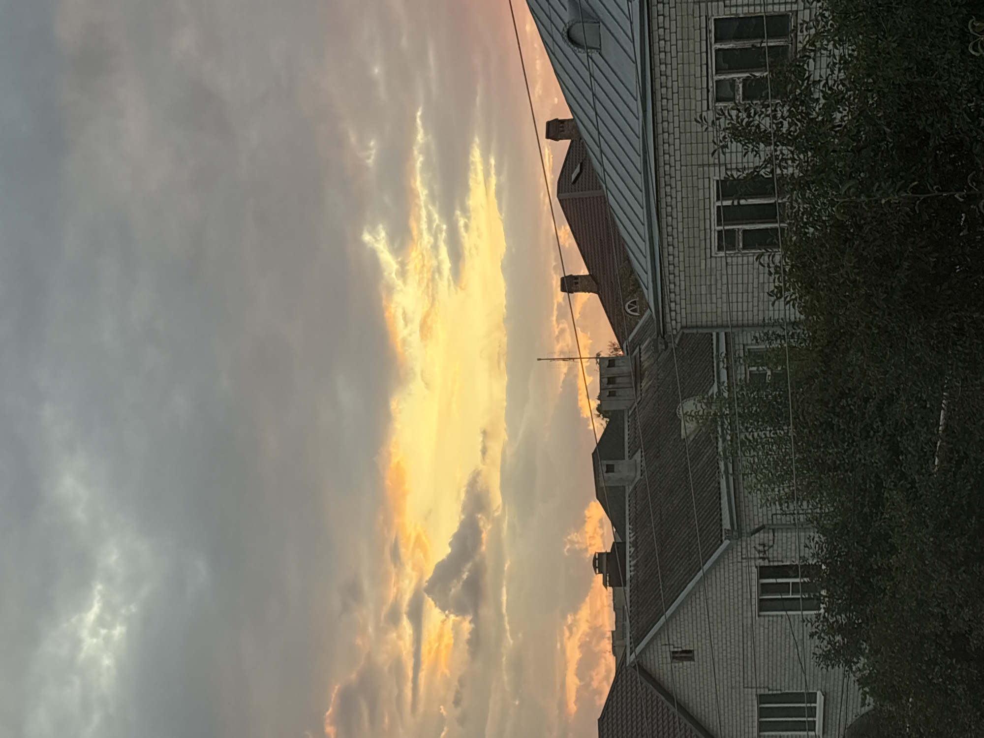 The image depicts a sky filled with dramatic clouds, illuminated by the warm hues of sunset, casting a golden and pink glow. Below the sky, the rooftops of several buildings are visible. These buildings appear to be residential, with slate and metal roofing. Some have chimneys, and architectural details like dormer windows can be seen. The lower portion of the image shows the facades of some of these buildings, constructed from light-colored brick, with visible windows and the dark foliage of trees in the foreground. The scene conveys a serene, end-of-day atmosphere, likely captured from a vantage point overlooking a residential area in the city of Georgiyevsk, Russia.