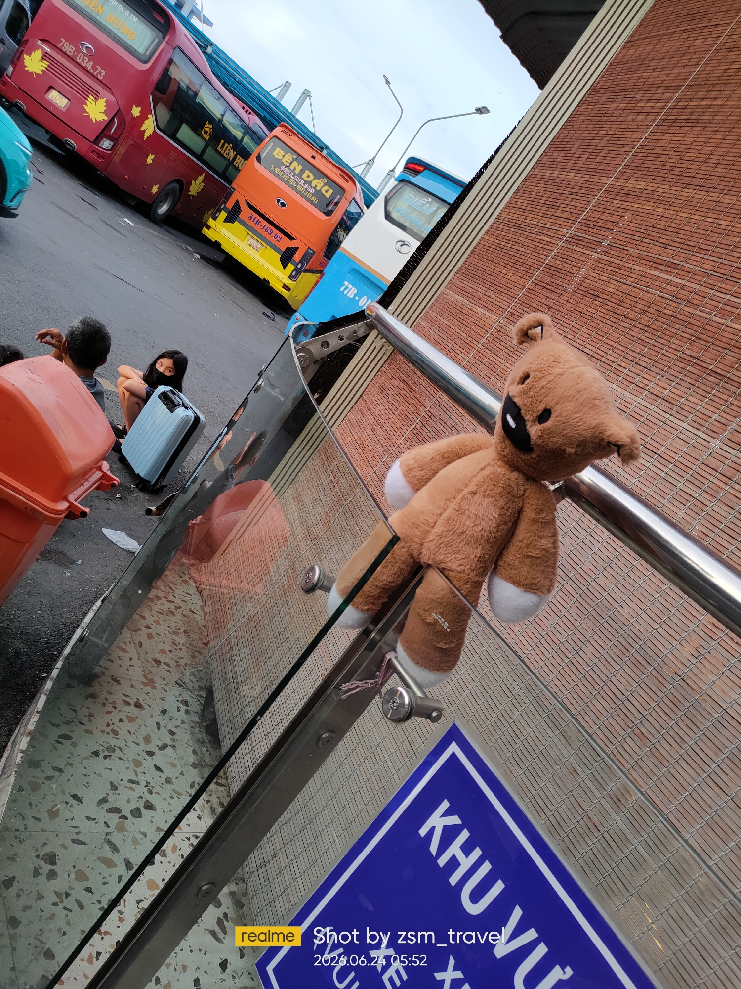 A teddy bear is hanging by its arm from a metal railing on an elevated walkway in Ho Chi Minh City, Vietnam. The walkway has a textured, brick-like wall on one side and a glass barrier on the other, revealing a street scene below. In the background, several large buses, a turquoise car, and people waiting with luggage are visible. A blue sign with white Vietnamese text is partially visible at the bottom right of the frame, indicating a "ZONE" or "AREA." The overall scene suggests a bus station or transportation hub.
