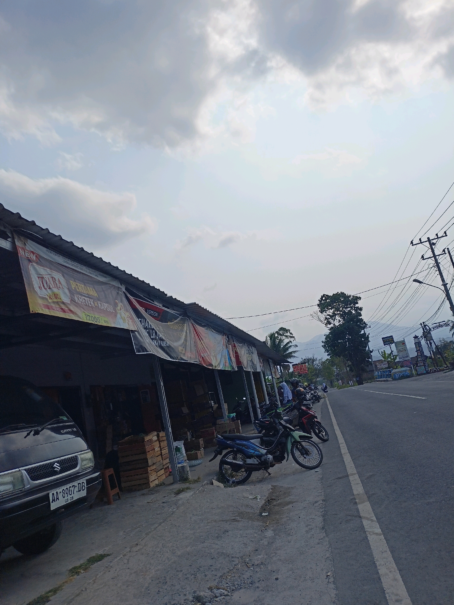 The image depicts a roadside scene in the city of Payaman, Indonesia, under an overcast sky. On the left, a small shop or market stall features a corrugated metal roof and is adorned with several large banners displaying text in Indonesian, one of which advertises "JUARA PERTAMA KRETEK". Stacks of wooden crates are visible behind the banners. In front of the stall, a black Suzuki Carry minivan is parked, with its license plate reading "AA 8907 DB". Several motorcycles are lined up along the edge of the road.
The road itself is paved and runs parallel to the row of shops. Power lines and utility poles are prominent on the right side of the road. In the background, mountains can be seen in the distance, partially obscured by trees. The overall atmosphere suggests a quiet, possibly rural or semi-urban, commercial area. No specific human activities or interactions are clearly discernible, beyond the presence of parked vehicles and potential occupants within the shaded area of the shop.