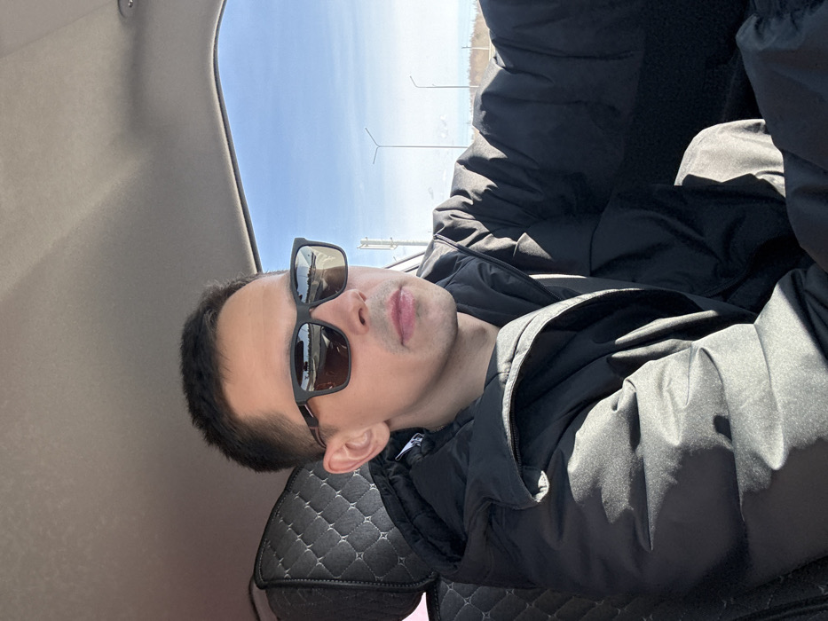 A male individual, appearing to be in his 20s or 30s, is seated in the driver's position of a vehicle in Kukushtan, Russia, looking forward and slightly to his right. He has short, dark hair and is wearing dark-framed rectangular sunglasses, which reflect a segment of a road with visible markings and distant vehicles or structures. He is dressed in a black puffer jacket over a dark undergarment. Strong direct sunlight illuminates his left side, casting defined shadows on his jacket and within the car's interior. The vehicle's interior features a light-colored headliner and a dark, diamond-quilted seat cover on the seat behind him. Through the side window, a clear, bright blue sky is visible above a distant, sparsely vegetated landscape containing several tall utility or light poles. The scene suggests a clear, sunny daytime environment on an open road.
