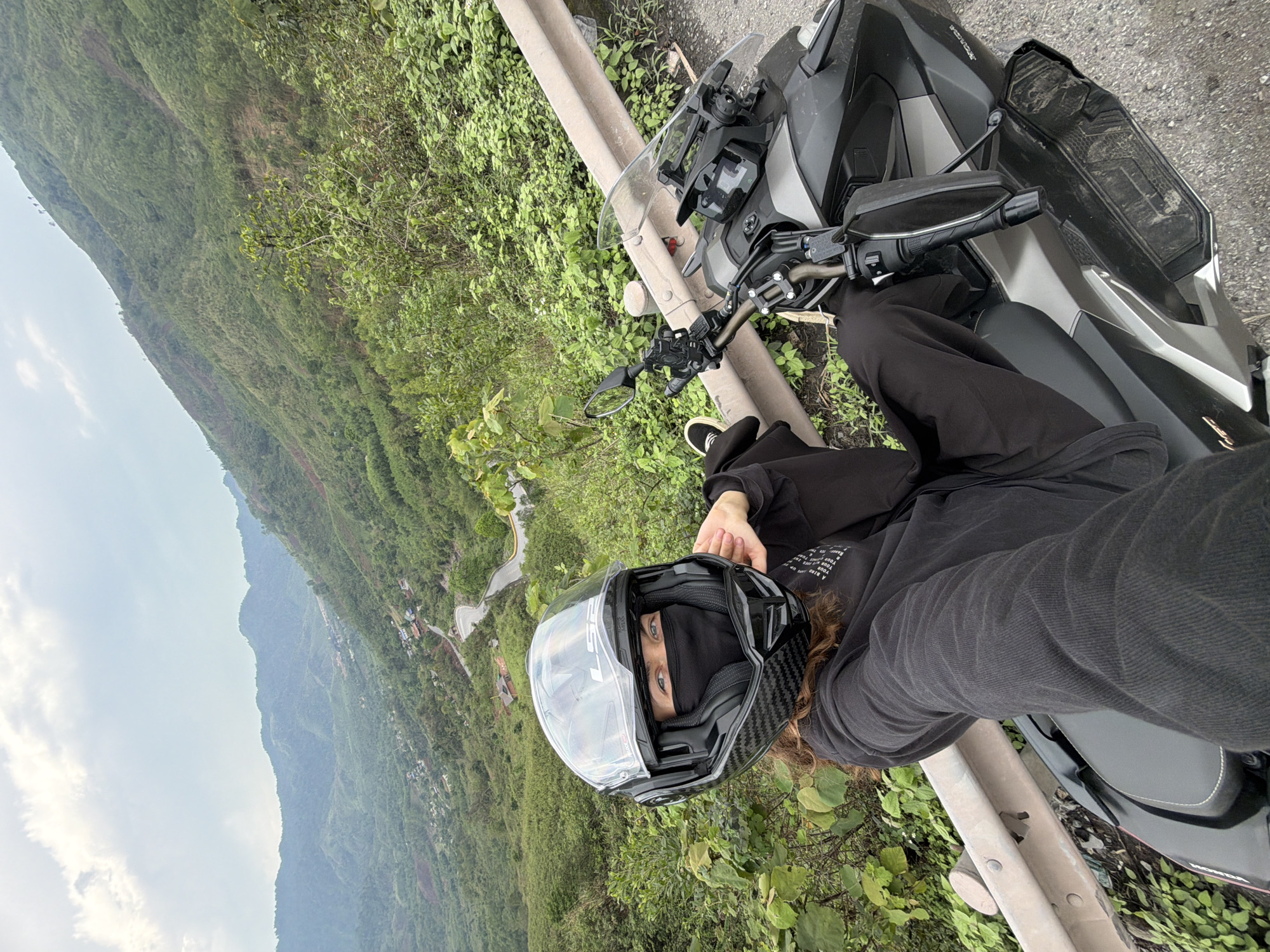 A person wearing a black helmet and dark clothing is positioned on a motorcycle adjacent to a guardrail. The motorcycle is parked on the side of a paved road with vegetation growing nearby. In the background, a mountainous landscape covered in lush greenery is visible. A road winds along the side of the mountain. The photo was taken in the La Lay Commune, Vietnam.