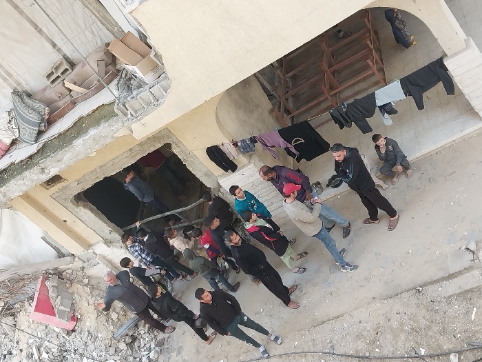 An aerial-perspective image depicts a group of approximately 20-25 males, ranging in age from children to adults, gathered in a damaged urban environment in Khan Yunis, Palestinian Territory. The scene features a partially destroyed or under-construction building made of light-colored concrete or stucco, with significant rubble, twisted rebar, and a red rectangular object visible in the upper left and next to a dark, rectangular doorway.
A line of individuals is entering or moving through this doorway, which leads into a darkened interior space. An older man in a grey shirt and dark pants stands to the left of this entrance, with both arms extended outwards, seemingly directing or observing the activity. Other individuals stand on the uneven, light-colored ground, some observing, others appearing to wait. One boy crouches on the far right, and another individual squats closer to the center.
Above the entrance on the damaged building structure, various items including blankets, cardboard boxes, and concrete blocks are visible. To the right, clothes are hanging on a line or rail, and a wooden frame structure is partially visible under an archway, suggesting further construction or debris. A black cable runs along the lower left foreground. The overall scene indicates a group of people engaged in an activity around a damaged structure.