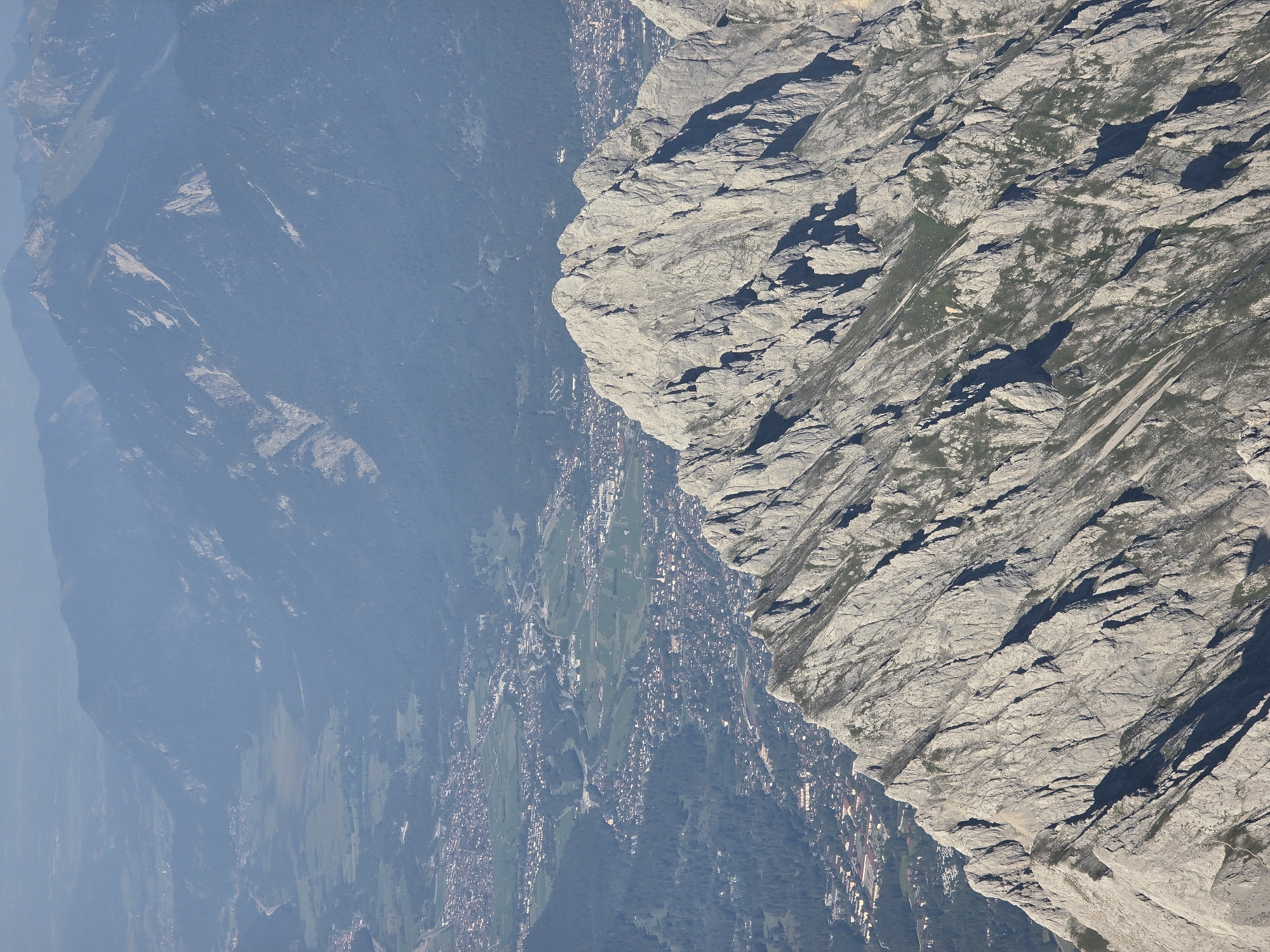 This is a high-angle aerial photograph of a mountainous region, likely the Bavarian Alps, with a town visible in the valley below. The foreground is dominated by a rugged, rocky mountain ridge characterized by sharp peaks, deep crevices, and patches of sparse green vegetation. The rock faces are predominantly grey and appear weathered.
In the middle ground and background, the terrain softens into rolling hills and larger, more distant mountain ranges. These further mountains are covered in dense, dark green forests, with some lighter green patches indicating meadows or cleared areas. The distant peaks are hazy and indistinct, suggesting a significant distance.
Nestled in the valley between the mountains is a town or city, identifiable by its densely packed buildings, roads, and cultivated fields. The buildings appear to be mostly low-rise with red-tiled roofs, typical of Bavarian architecture. A river or stream can be seen meandering through the valley, bordered by green fields. The overall impression is one of a vast, natural landscape encompassing a human settlement.
Based on the geography and the presence of the town, this image likely depicts a view towards Garmisch-Partenkirchen, Germany, from one of the surrounding mountain peaks such as the Zugspitze or the Alpspitz. The town's layout, with its proximity to forested mountains and its setting in a valley, is consistent with Garmisch-Partenkirchen. There are no people or discernible actions depicted in the image. The environment is natural and mountainous, with a human settlement integrated into the landscape.