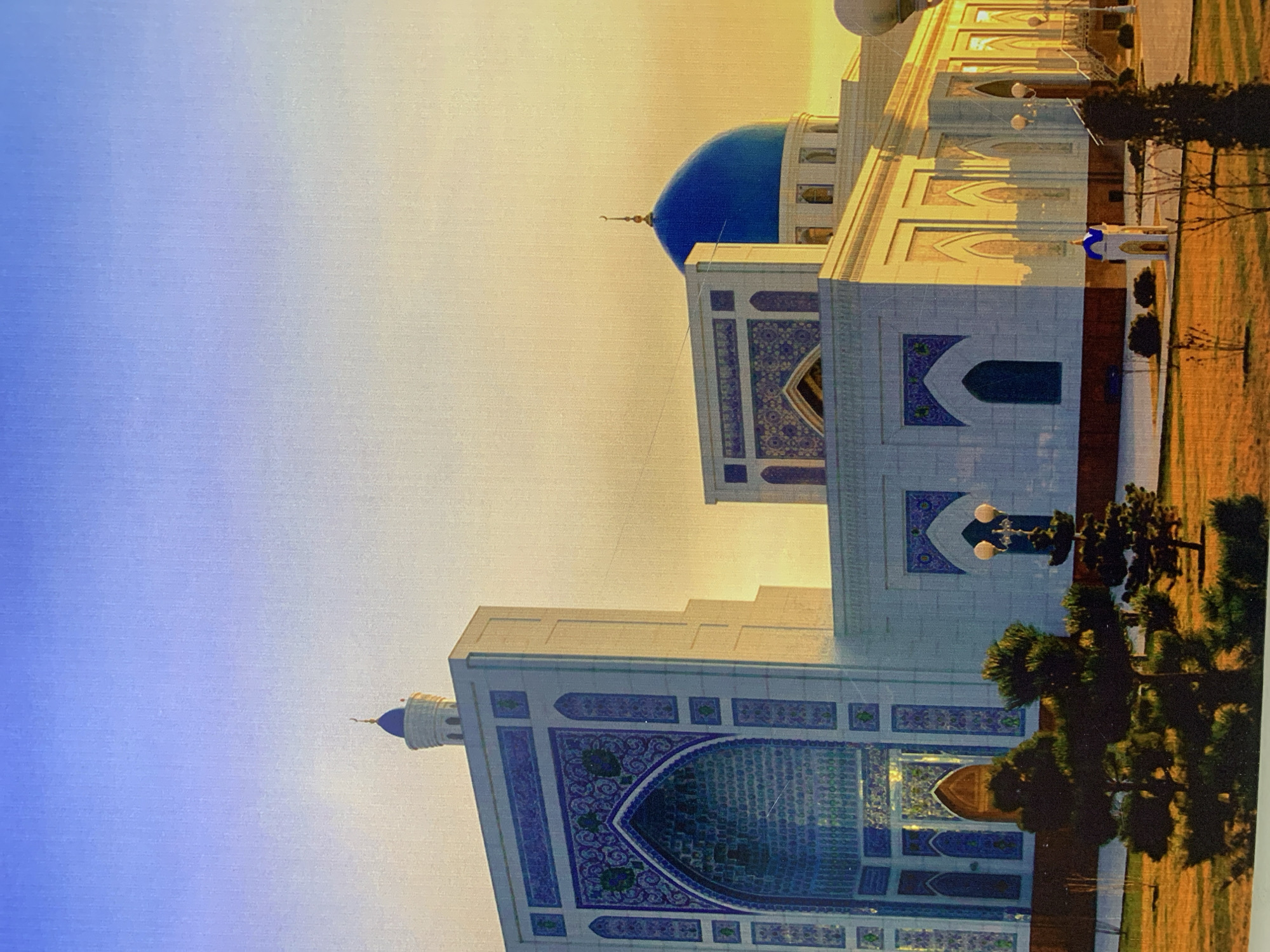 Here's a detailed description of the image:
The image shows a large, ornately decorated building with a prominent blue dome. The architectural style appears to be Islamic, with detailed geometric patterns and arched windows. The building's facade is predominantly white with blue and purple accents in the mosaic tilework. The building is situated in an open area with trees and grass. The sky transitions from blue to a warm yellow, indicating either sunrise or sunset. It is located in the city of Unknown, Unknown.