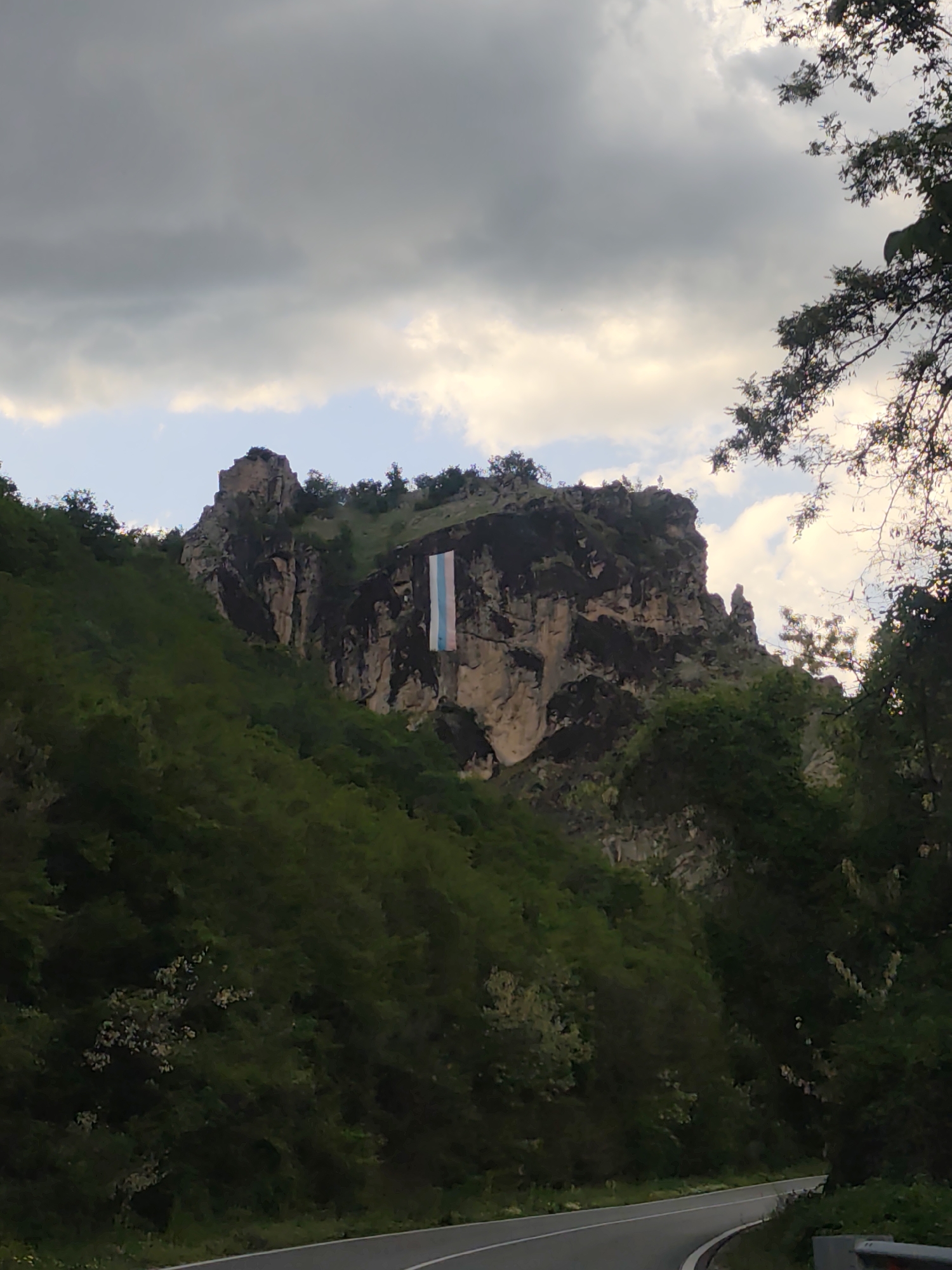A vertical photograph shows a road curving around a mountain in Bukovo, Bulgaria, with a trans flag hanging on the mountain’s rock face.
The mountain is rocky and covered with green vegetation. A large trans flag hangs prominently from the face of the rock. The road is paved and curves through the lush green landscape. Overhanging trees and bushes obscure part of the road. The sky is overcast with heavy, dark clouds. The upper right corner shows a tree with dark, leafy branches.