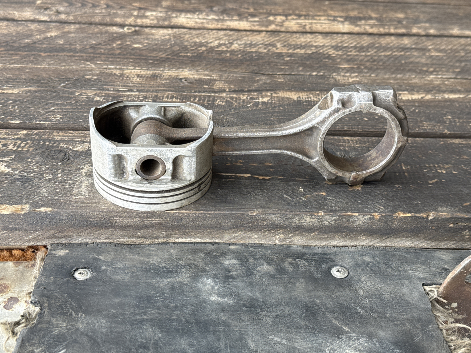 The image depicts a used piston and connecting rod resting on a weathered wooden surface. The piston shows signs of wear, with scoring marks and discoloration. The connecting rod appears intact, although also displaying oxidation. The background consists of dark, aged wood with visible grain. The lower portion of the image reveals a contrasting dark gray or black material, possibly another section of the wooden surface, fastened with screws. The image was taken somewhere in the world, but there is no support to confirm that the image comes from the city of Perm, Russia.