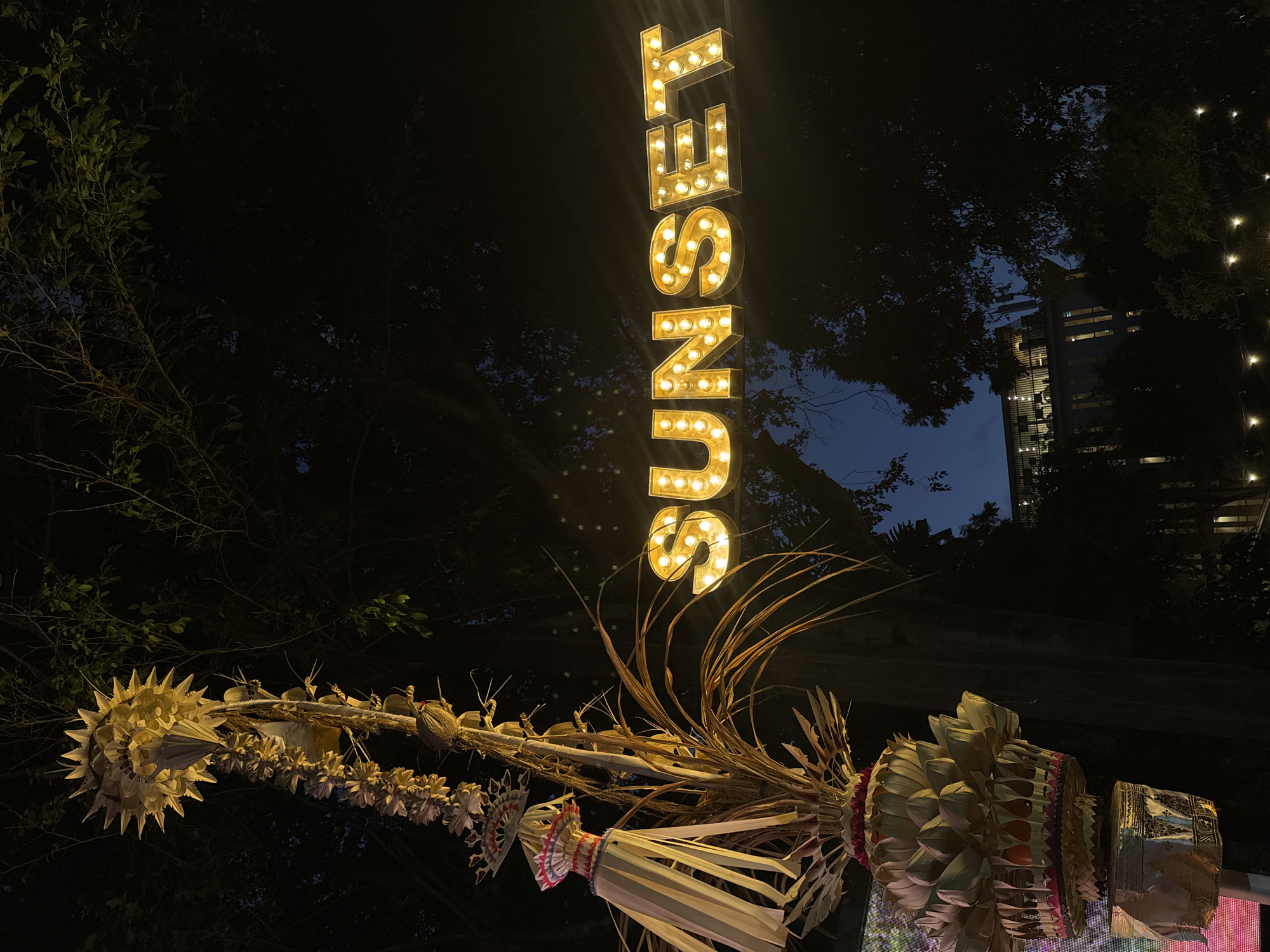 A vertical sign with illuminated letters spells out "SUNSET." The letters are gold with exposed bulbs that glow warmly. Below the sign, a decorative arrangement of dried palm leaves and woven materials hangs, possibly a traditional Balinese offering. The scene is set at night, with the dark sky and silhouettes of trees in the background. In the distance, a modern building with illuminated windows is visible, suggesting an urban or semi-urban setting. The overall mood is festive and possibly a bit mysterious due to the night time setting. The location is identified as Kuta, Indonesia.