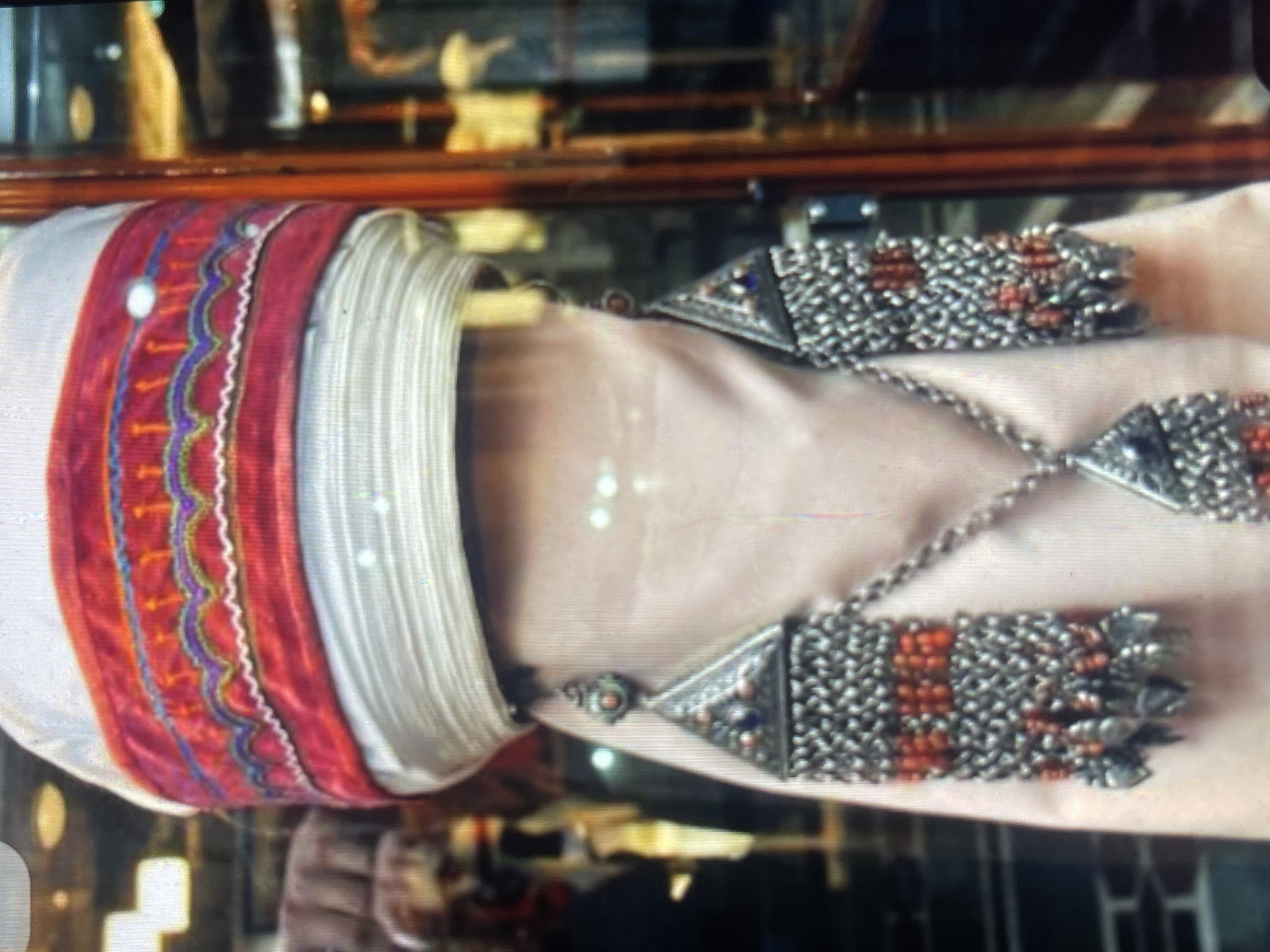 A close-up shot of a mannequin displaying traditional Kyrgyz attire, specifically a part of a headpiece and ornate jewelry. The headpiece is a tall, wrapped turban-like structure, predominantly white with horizontal ribbed textures. A wide band of red fabric is wrapped around the middle of the headpiece, embellished with intricate zigzag patterns in yellow, purple, and blue thread, with small white circular accents.
Suspended from the sides of the headpiece, near where ears would be, are large, triangular earrings. These earrings are made of silver-toned metal and feature a decorative triangular top with inset colorful stones, possibly blue and red. Dangling from the triangular tops are chains that form rectangular panels, intricately woven and filled with rows of small orange beads. More silver charms, resembling small leaves or teardrops, hang from the bottom of these rectangular panels. A central chain connects the two earrings, leading down to a larger, pendant-like piece that mirrors the design of the earrings, with a triangular top and a dangling decorated rectangular section.
The mannequin is dressed in a pale pink fabric that drapes downwards. The background is blurred and appears to be a display case or shop interior, with reflections of lights and possibly other items for sale. The scene suggests a cultural display or retail environment. The exact location in Bishkek City, Kyrgyzstan cannot be determined from the visual information provided in the image.