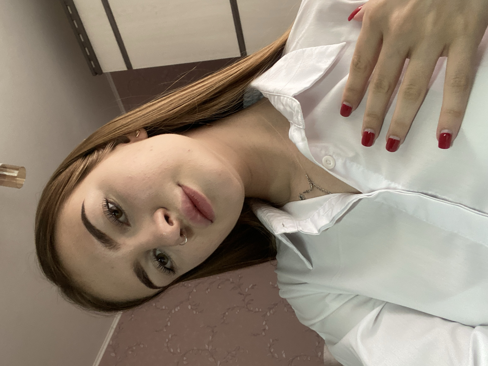 A young woman with long, brown hair, wearing a white button-down shirt and a necklace, is captured in a close-up, waist-up shot. She has a nose ring and her nails are painted red. Her right hand is placed on her chest, with her fingers spread. The background features a textured, light pink wall and a section of a white cabinet. The lighting suggests an indoor setting. The image is taken from a low angle, looking up at the woman.