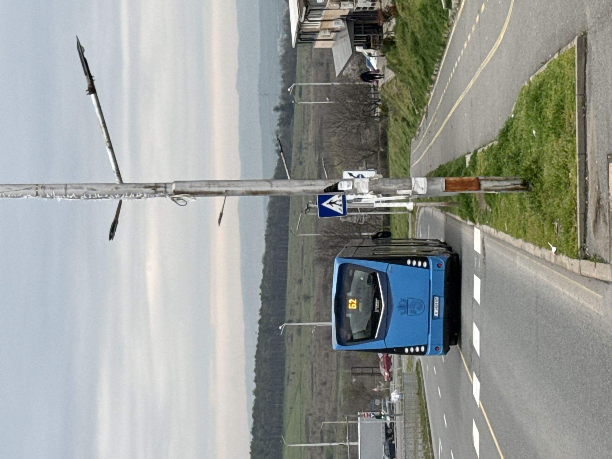 The image captures a blue city bus, displaying the number "52", navigating a road in Burgas, Bulgaria. The bus is positioned on the right side of the road, moving towards the camera. A pedestrian crossing sign is visible near the bus. The road is marked with white dashed lines and a solid yellow line. A sidewalk runs alongside the road, with green grassy areas bordering it. In the background, a landscape of trees and hills stretches out. A utility pole with a metal fixture is prominently featured in the foreground, partially obscuring the sky. The sky is overcast, suggesting diffused lighting. Buildings and other infrastructure can be seen further in the background, indicating an urban or suburban setting.