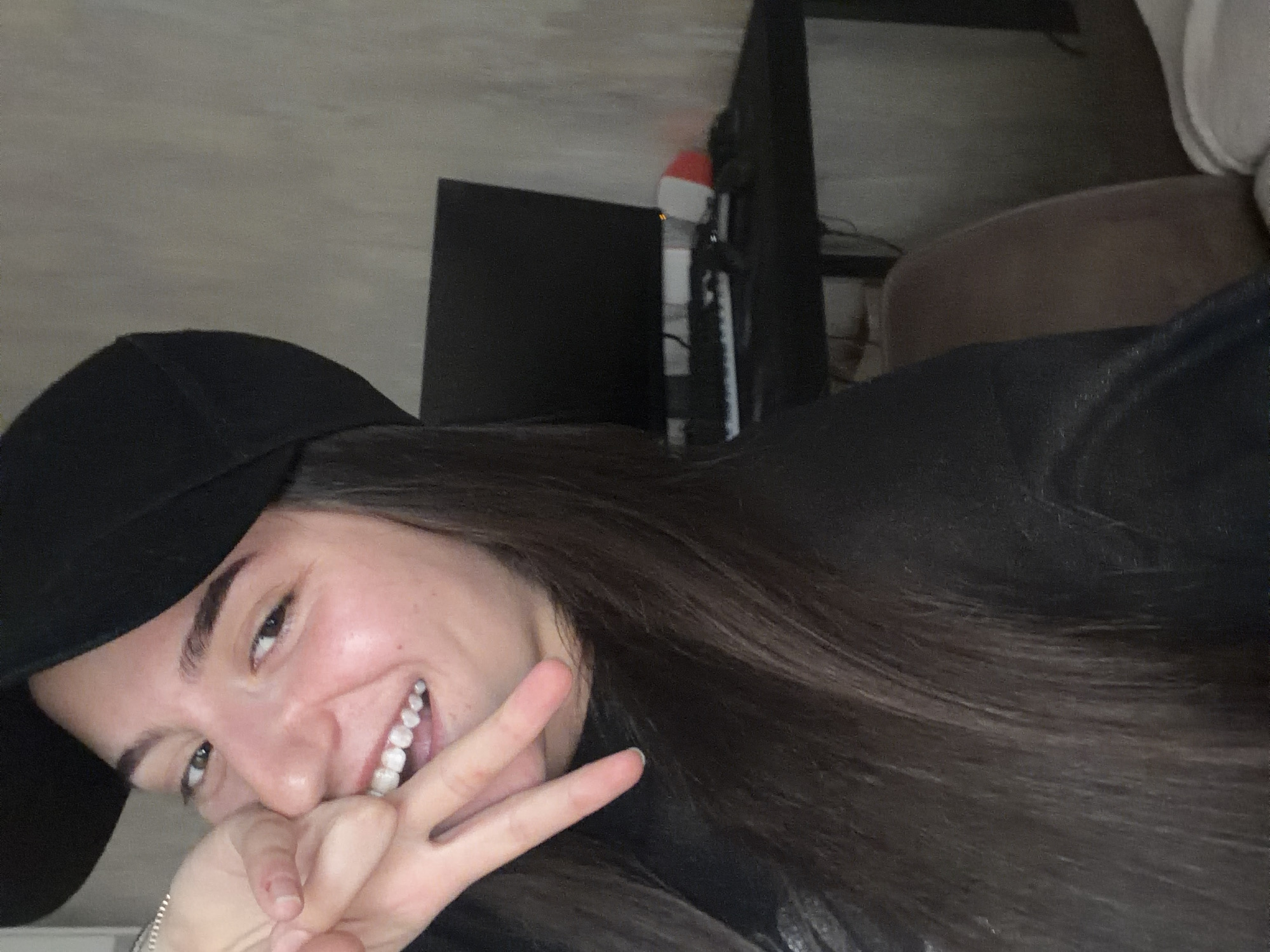 The image is a close-up selfie of a young woman with dark hair, wearing a black baseball cap and a black top. She is smiling and making a peace sign with her fingers near her mouth. Her left hand is visible, with a bracelet on her wrist. In the background, a desk with a computer monitor, a keyboard, and some electronic equipment is visible. The setting appears to be indoors, possibly a room. There is no information in the image that suggests a specific location in the city of Burgas, Bulgaria.
